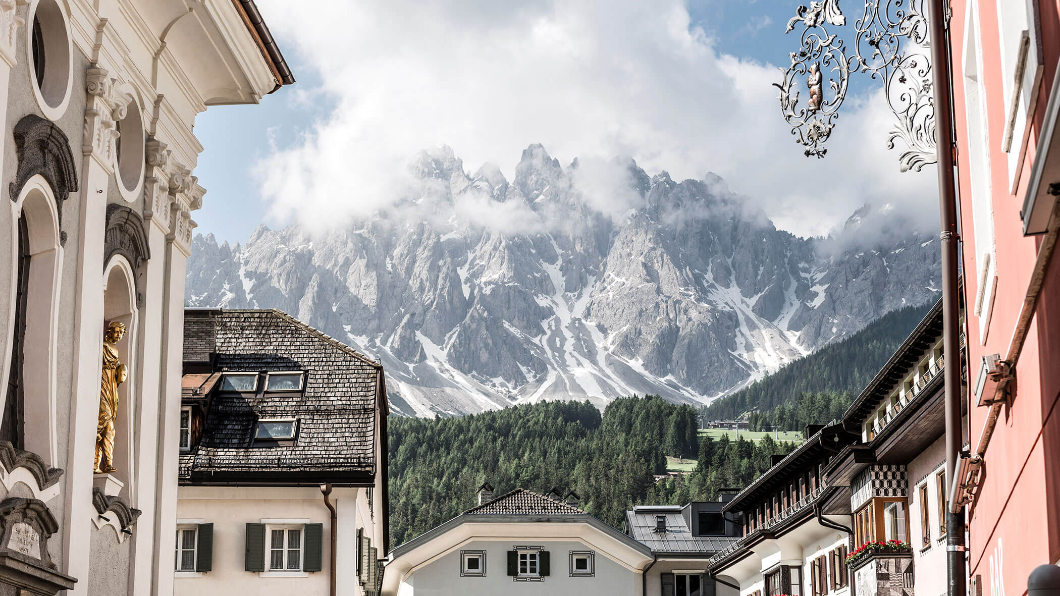 Snowy mountains rise behind quaint European buildings and trees under a partly cloudy sky.