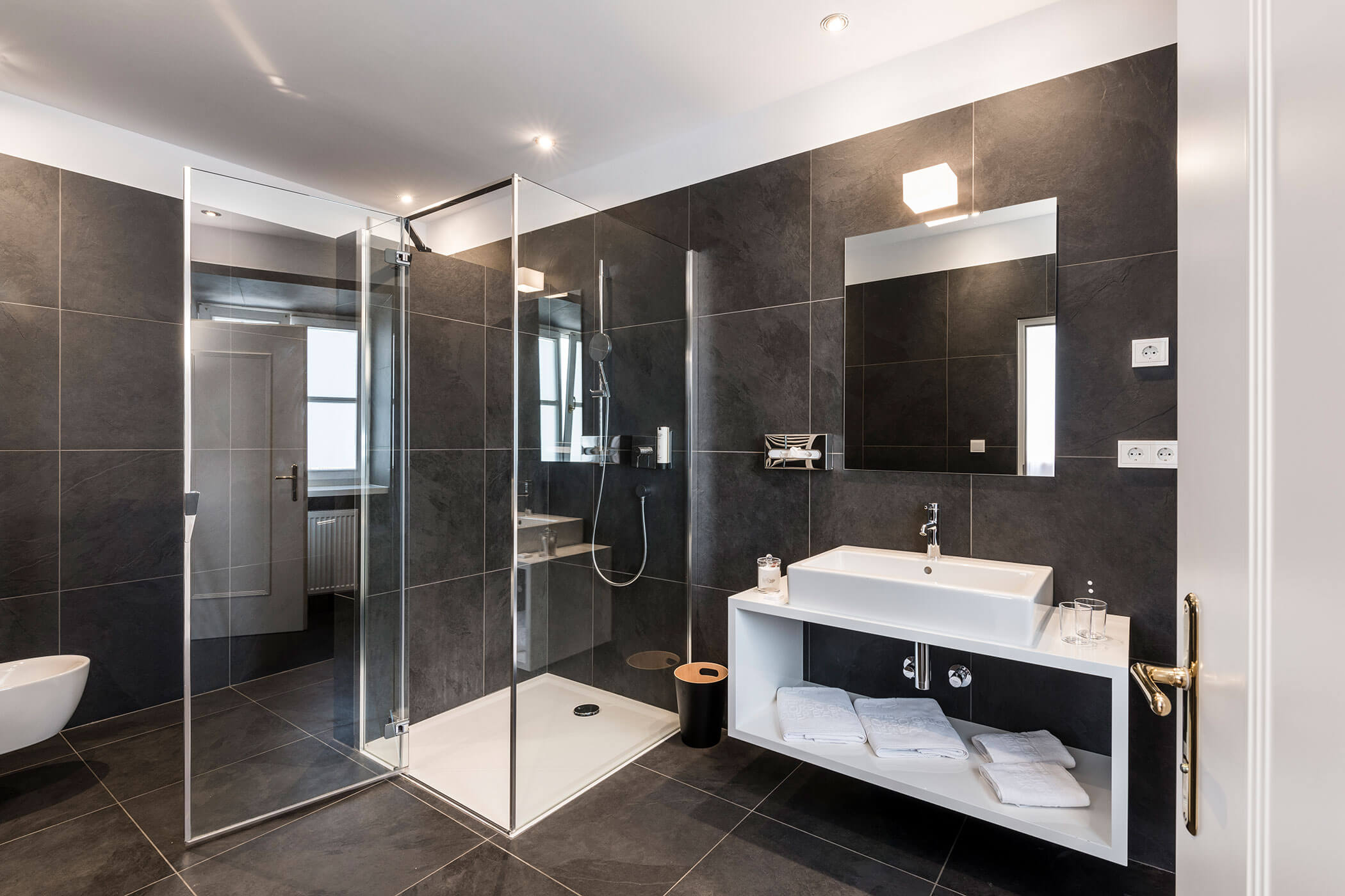 Modern bathroom with a glass shower, white sink, large mirror, dark tile walls, and neatly folded towels.