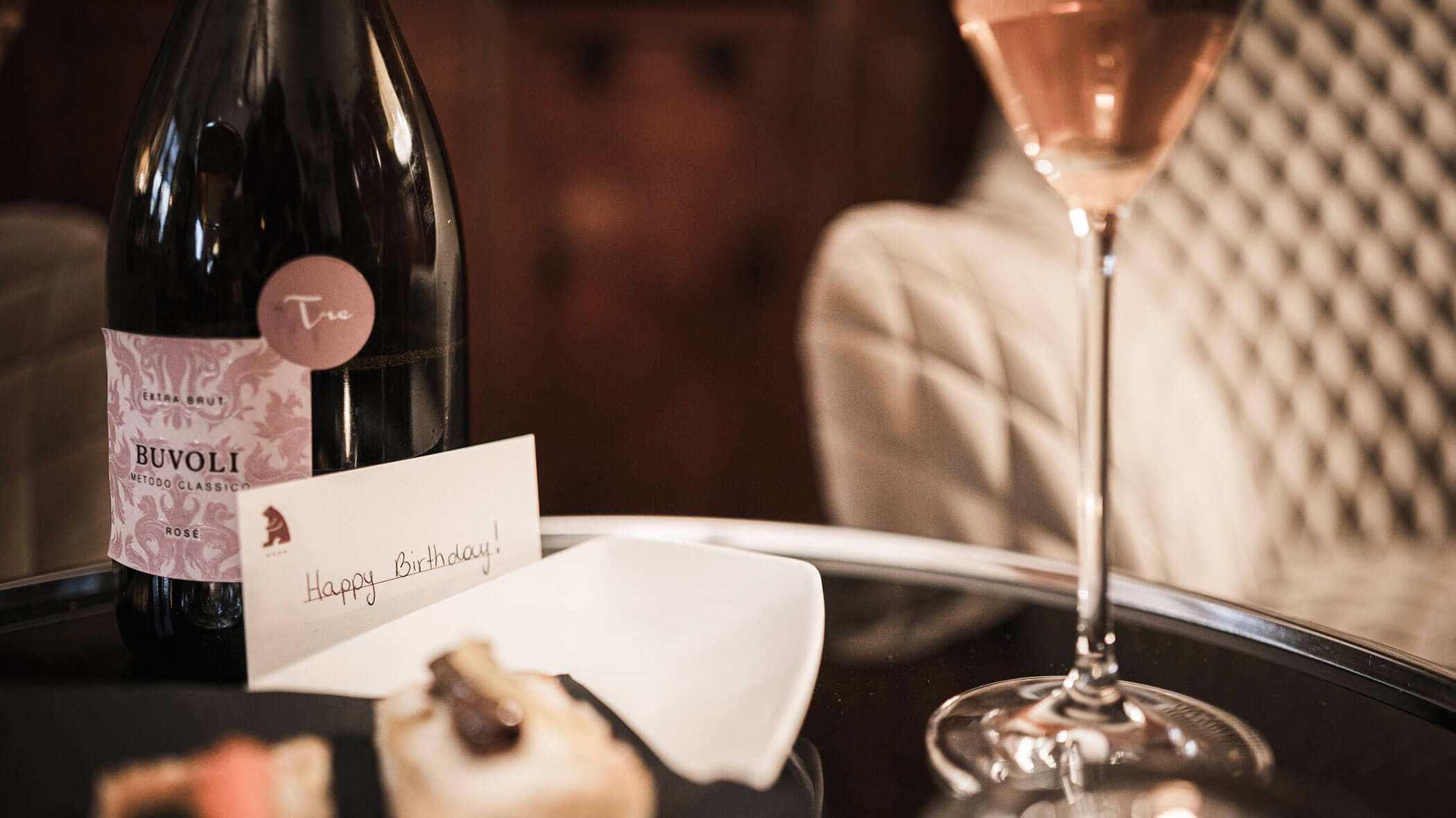 A bottle of sparkling rosé, a glass of wine, pastries, and a card reading Happy Birthday on a table.