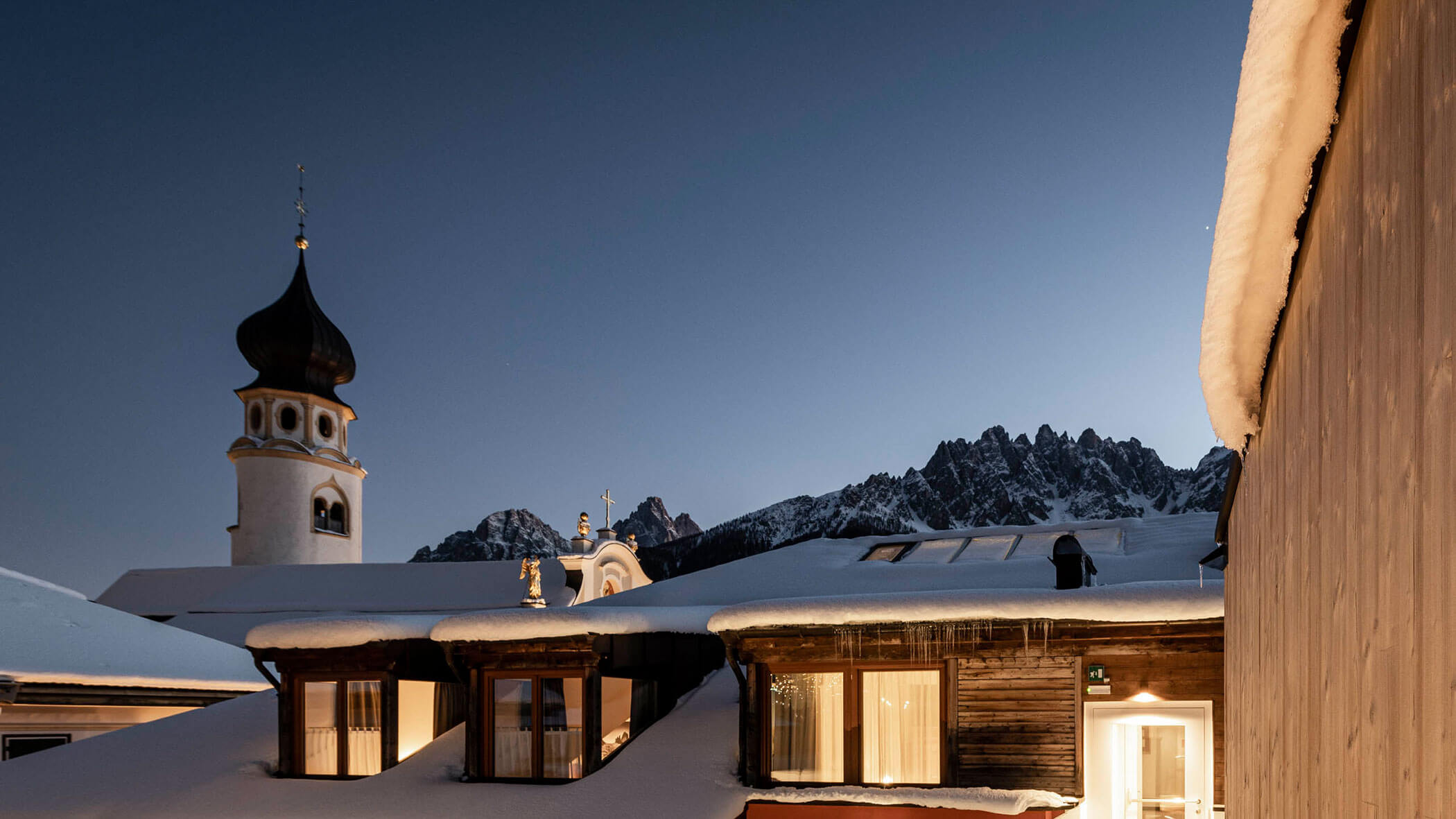 Edifici alpini innevati e una chiesa al tramonto, con le montagne sullo sfondo in un cielo limpido.