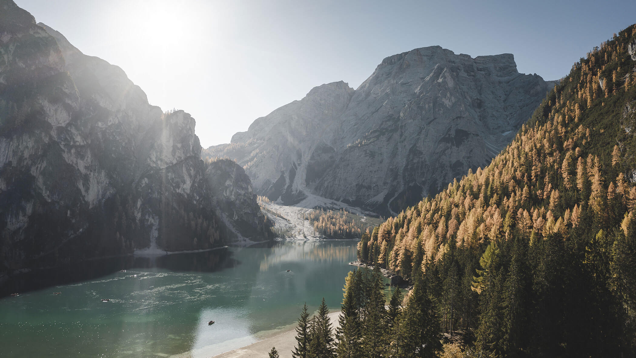 Mountain lake surrounded by pine trees, with sunlight shining over tall rocky peaks in the background.