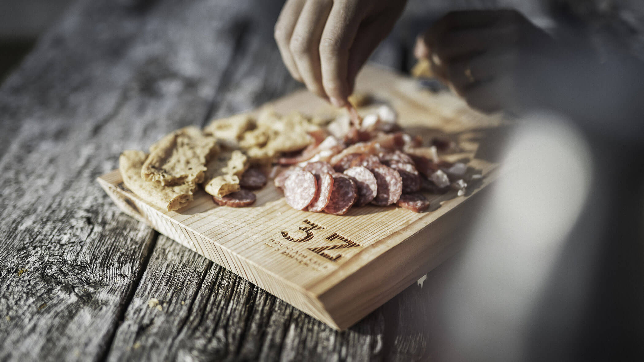 A hand arranges sliced salami and bread on a wooden board atop a rustic table.