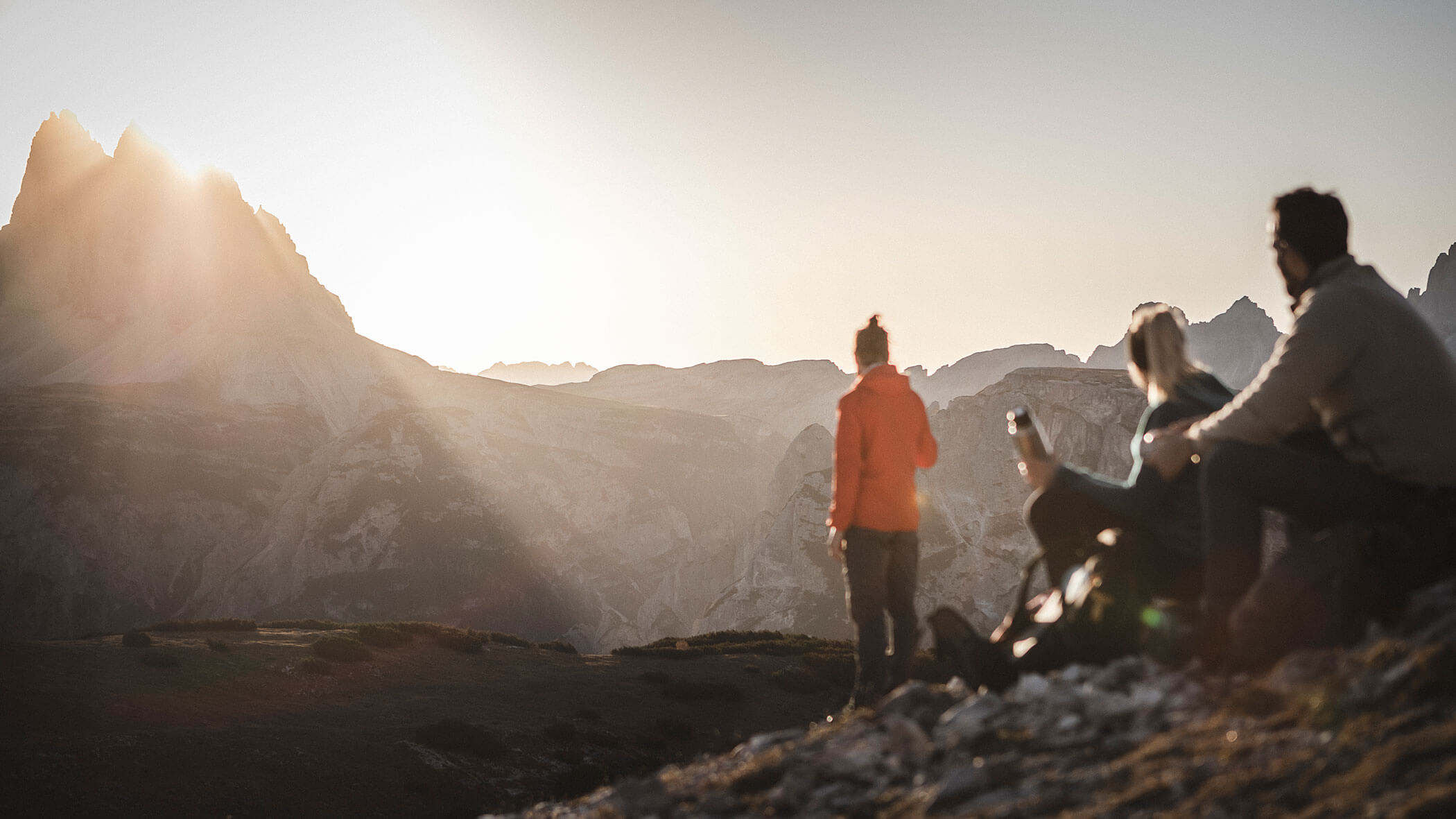 Three people enjoy a scenic mountain view at sunset, with sunlight streaming over rocky peaks in the distance.