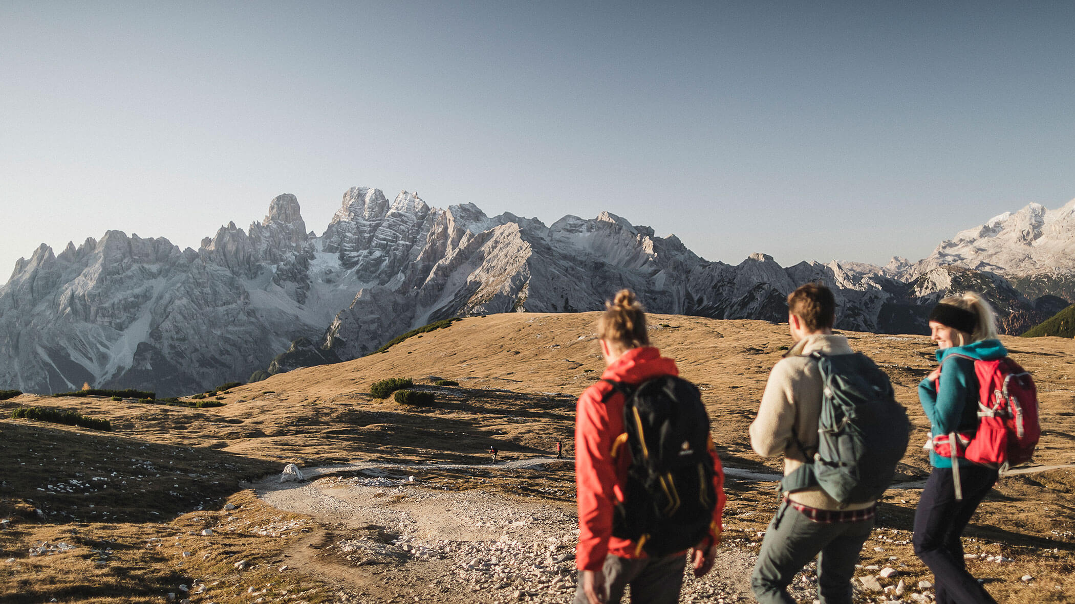 Three hikers with backpacks walk along a rocky trail toward snowy mountains under a clear sky.