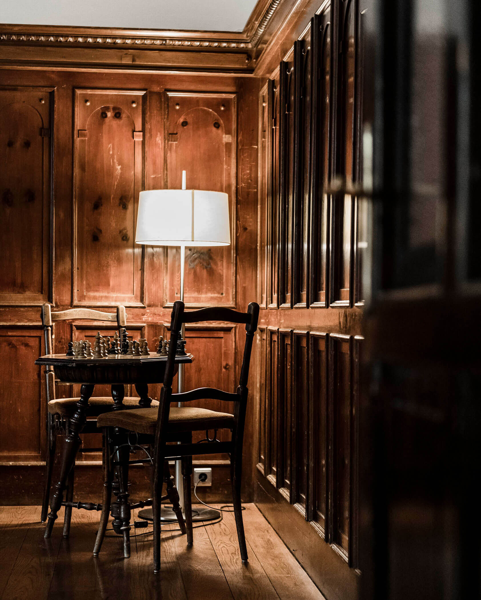 A cozy wooden room with a chessboard on a small table, two chairs, and a floor lamp.