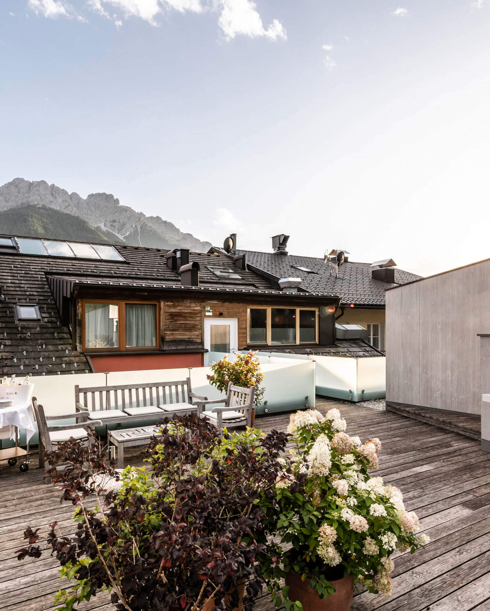 A rooftop patio with wooden decking, outdoor furniture, potted plants, and mountain views in the background.