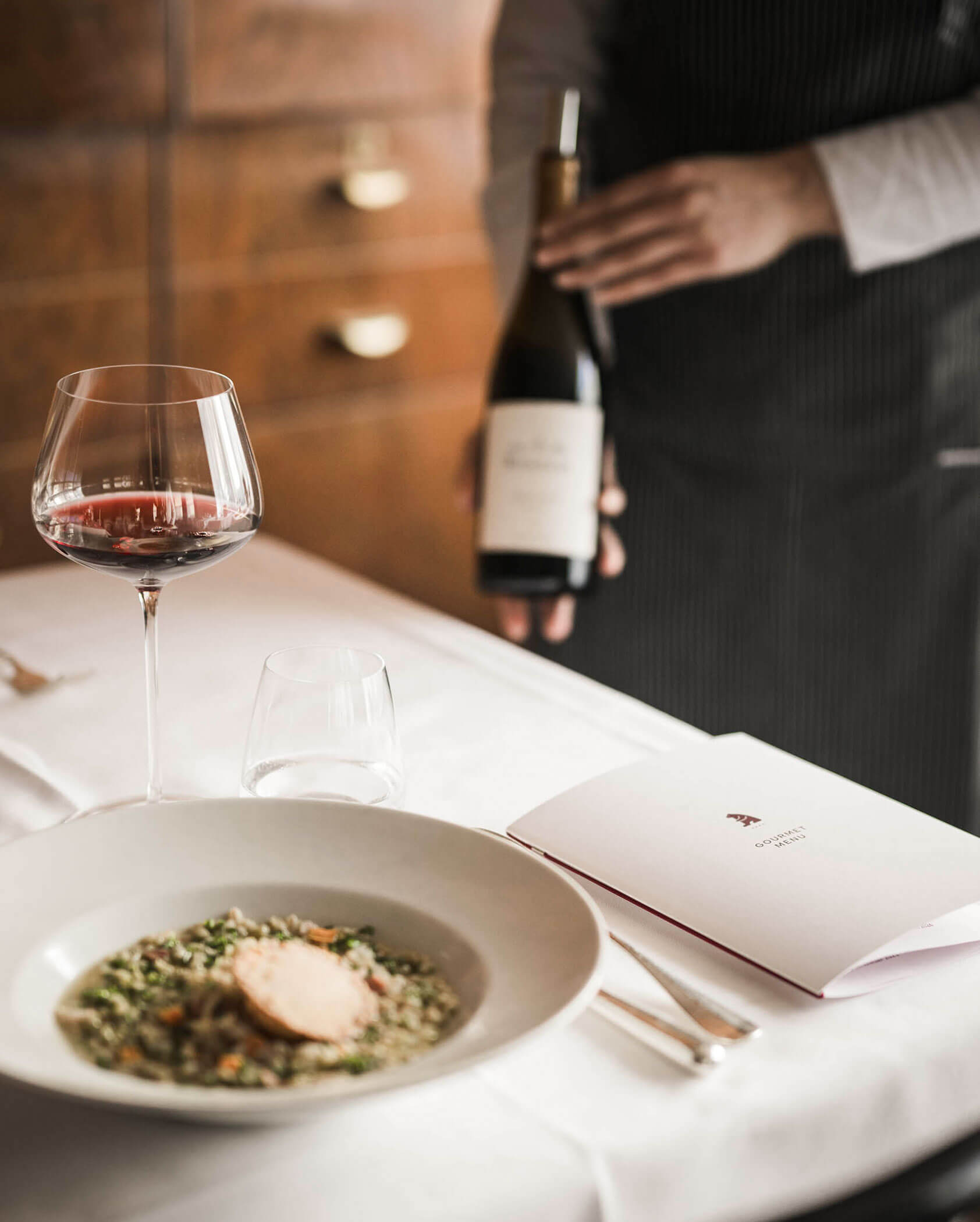 A waiter presents a bottle of wine next to a table set with risotto, wine, water, and a folded menu.