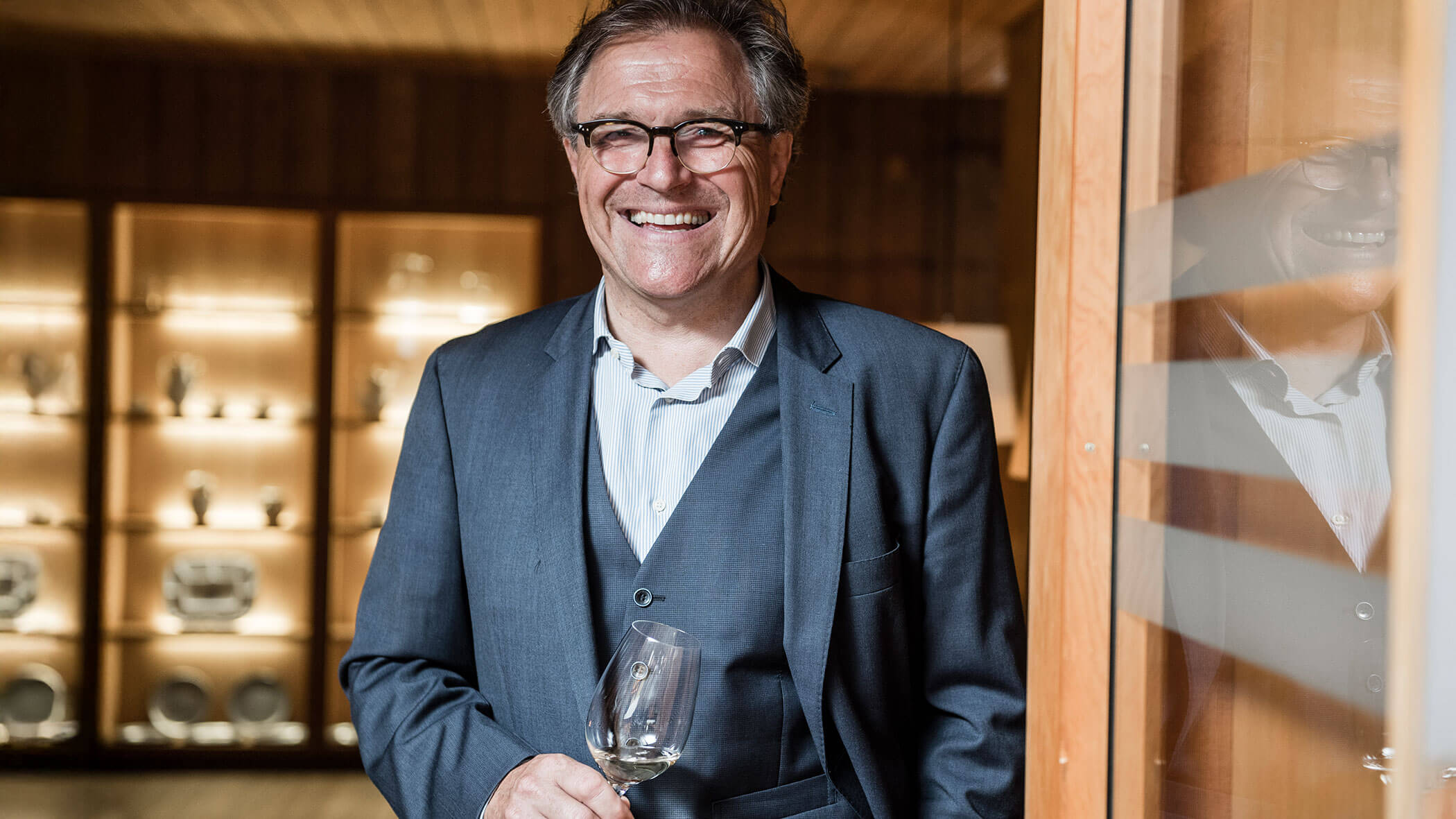 A smiling man in a suit holds a wine glass, standing in front of shelves with glassware.
