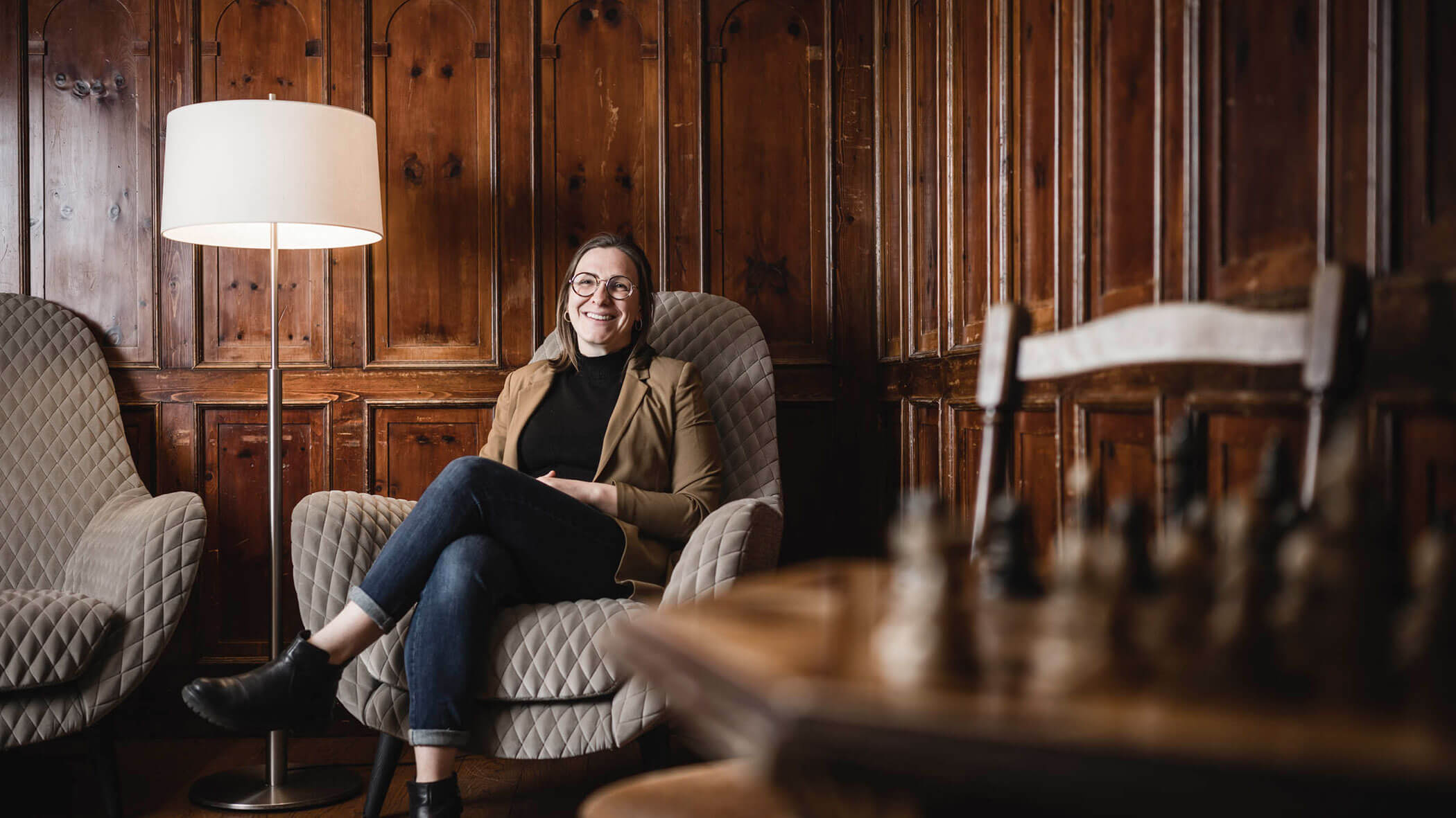 Smiling woman sits in an armchair by a lamp in a wood-paneled room, with a chessboard in the foreground.