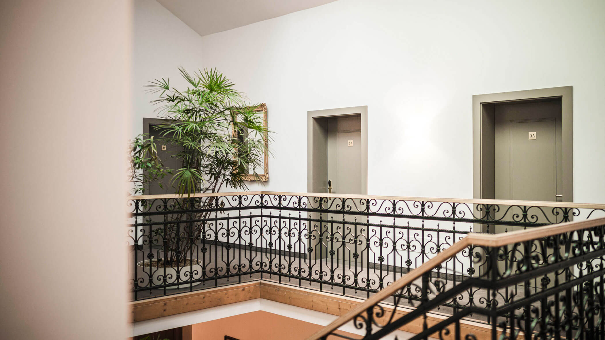 A staircase landing with ornate railing, three grey doors, a tall plant, and a framed mirror on the wall.