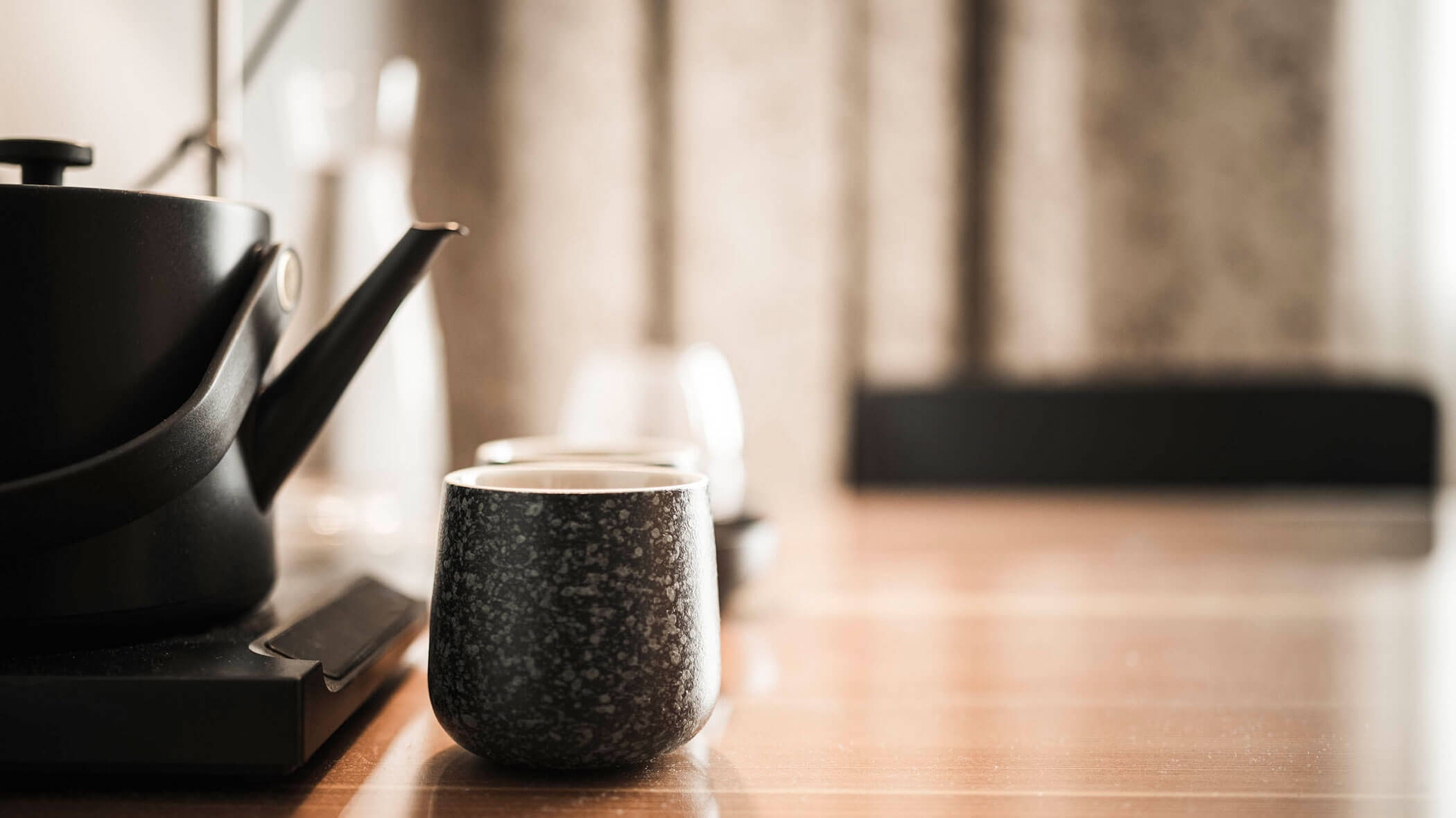 A black teapot and a dark ceramic cup sit on a wooden table with blurred background.