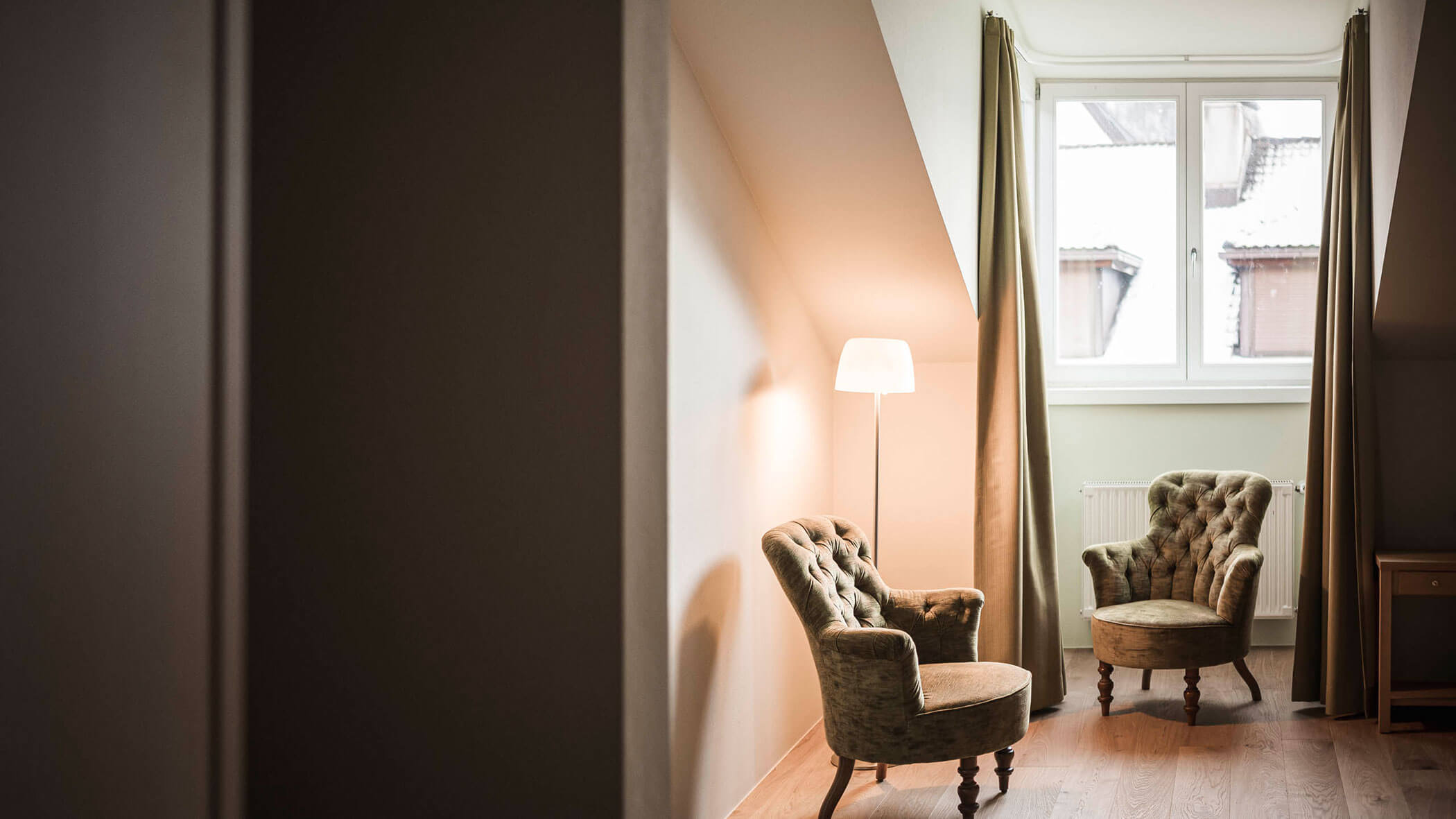 Two vintage armchairs and a floor lamp sit by a window with curtains in a cozy, softly lit room.