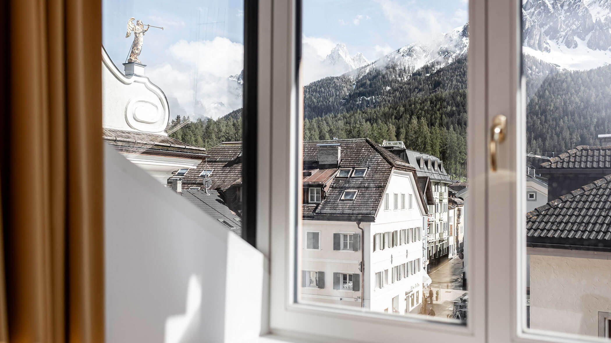 View through a window of alpine buildings, forested hills, and snow-capped mountains in the distance.