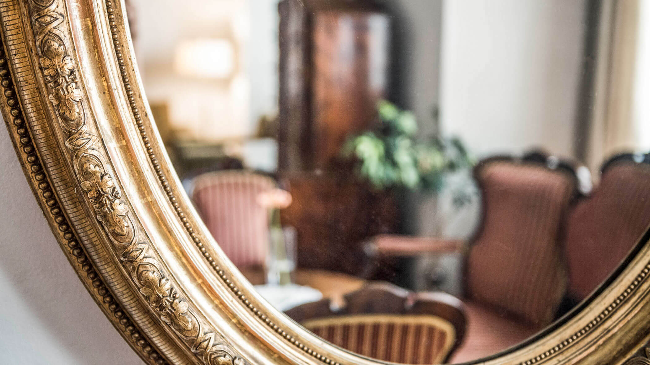 Close-up of an ornate gold mirror reflecting a cozy room with chairs and a plant.