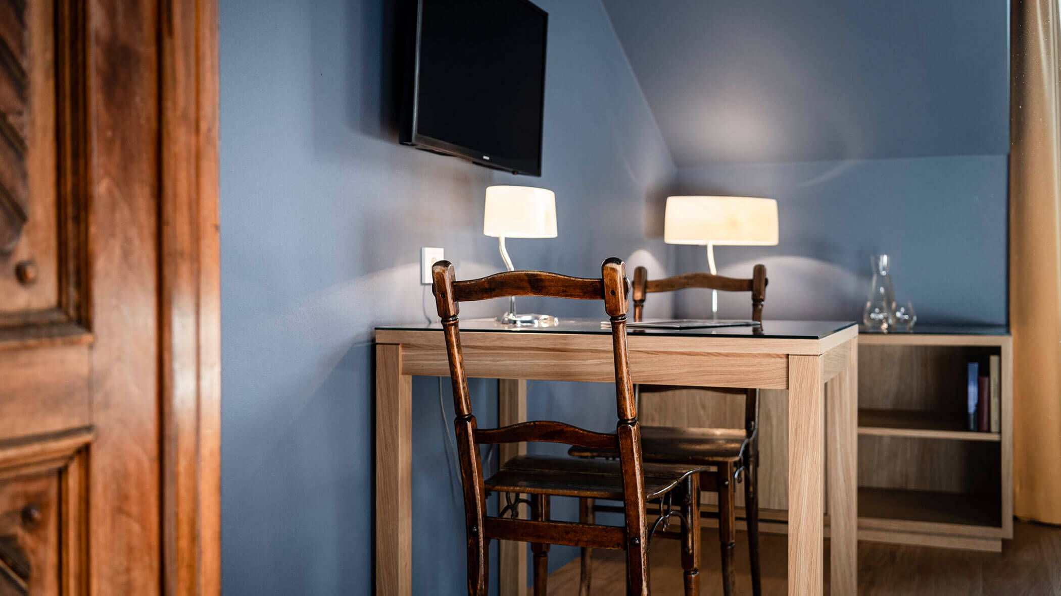 A wooden desk with two chairs, two lamps, and a wall-mounted TV in a blue-walled room.