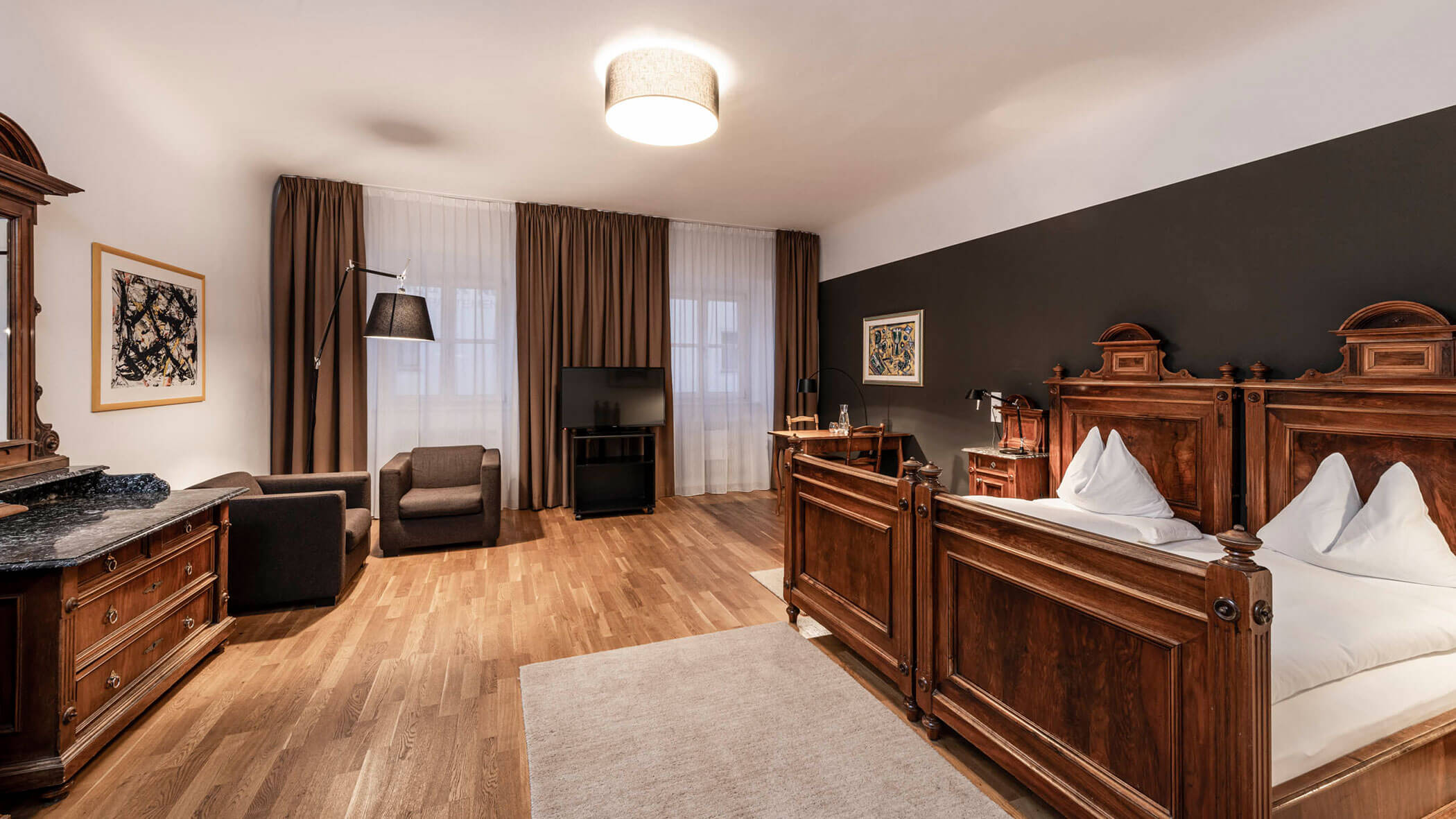 Spacious hotel room with vintage wooden furniture, a double bed, armchairs, and large windows with brown curtains.