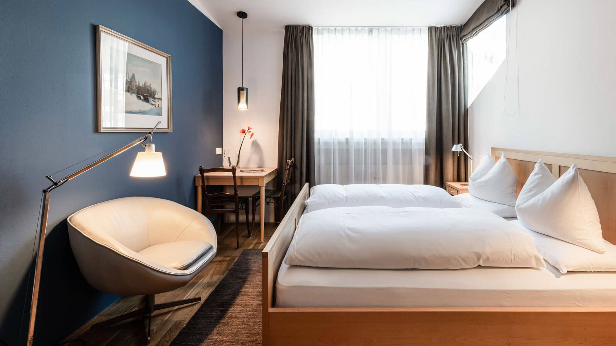 Modern hotel room with a double bed, armchair, desk, window with sheer curtains, and blue accent wall.