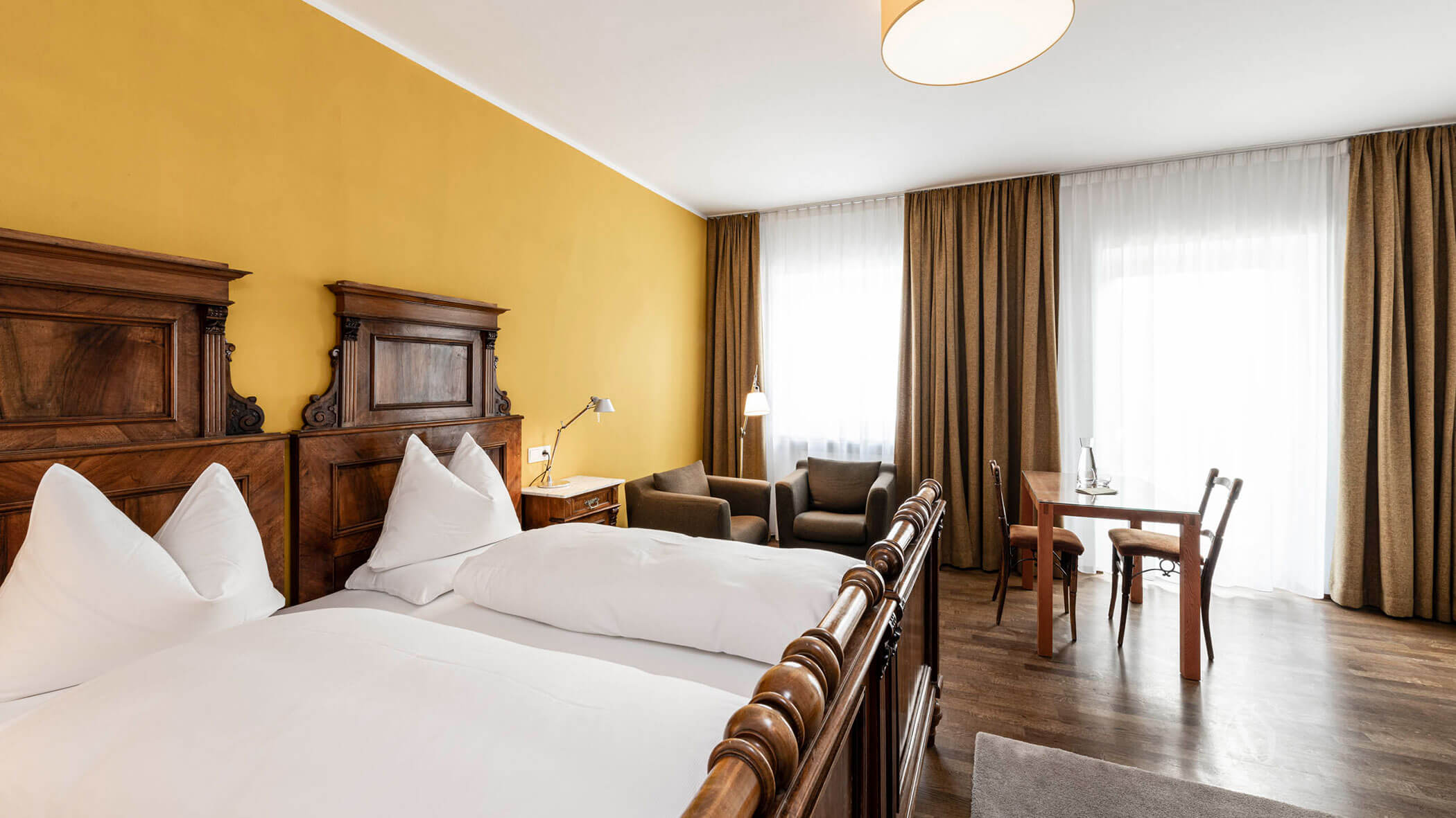 Bright hotel room with wooden bed, chairs, table, and large windows with brown curtains and white drapes.
