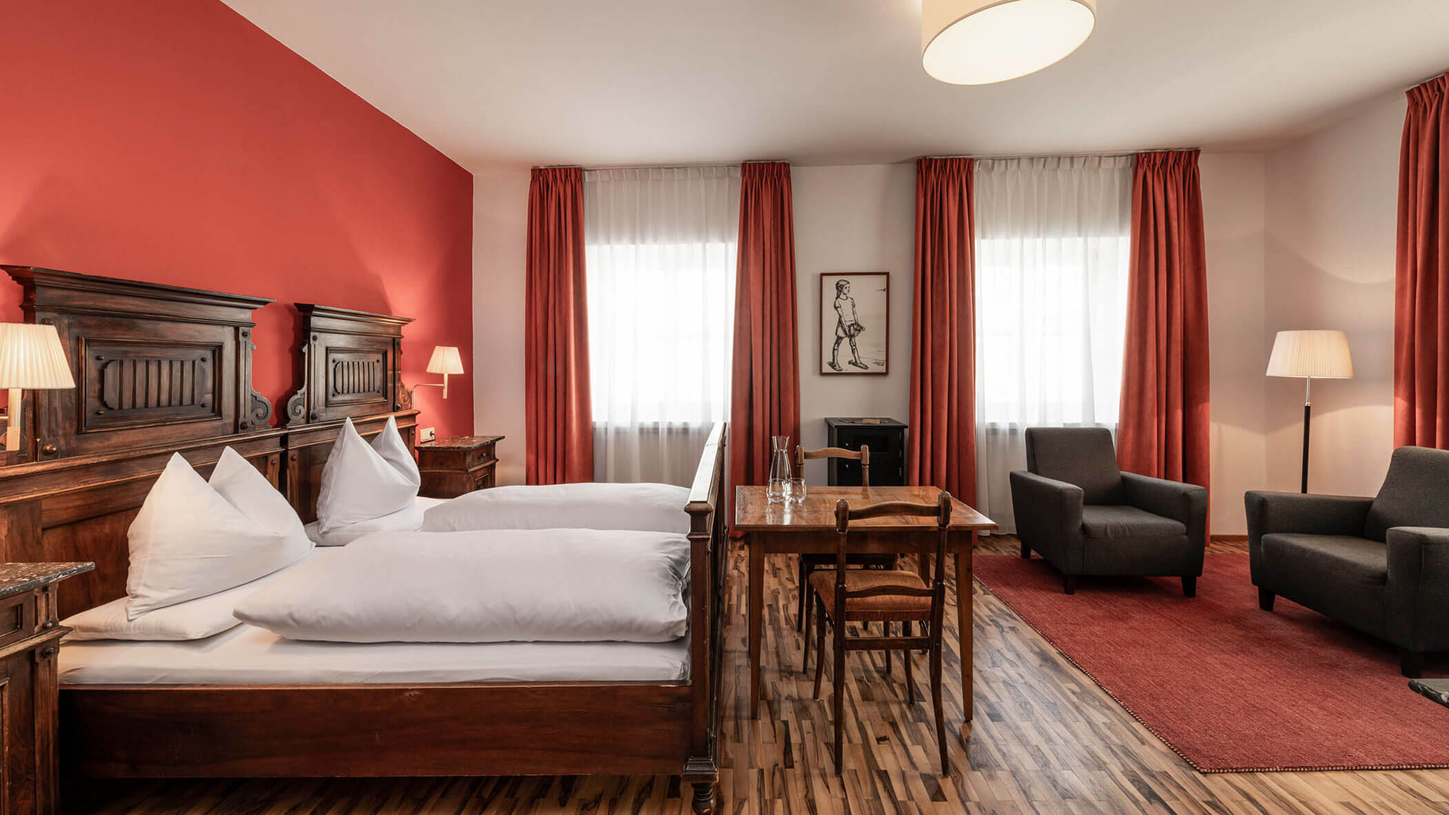 A cozy hotel room with a double bed, wooden furniture, red curtains, and a sitting area with two armchairs.