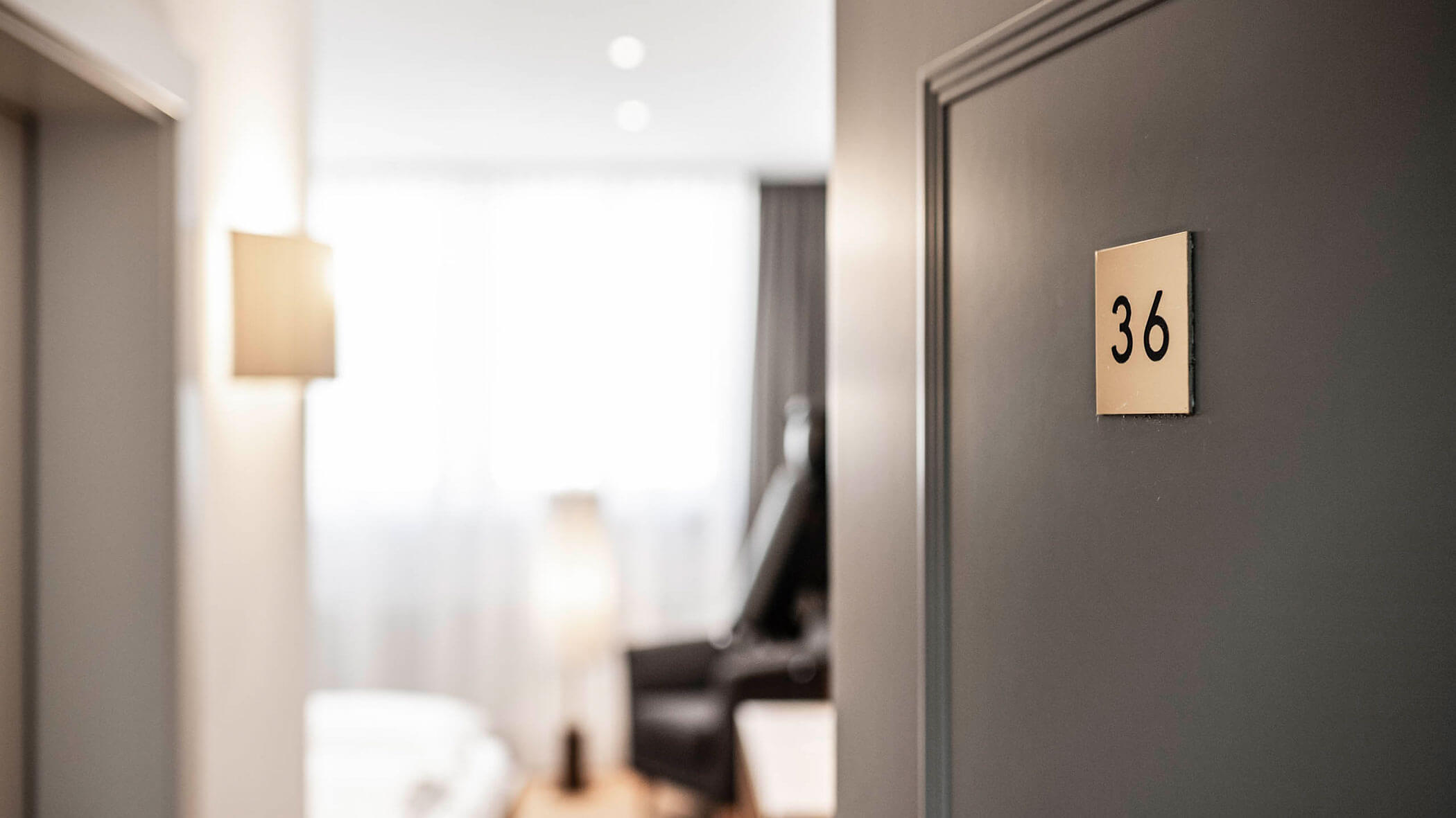 Gray door with a gold plaque showing the number 36, leading into a bright, modern hotel room.