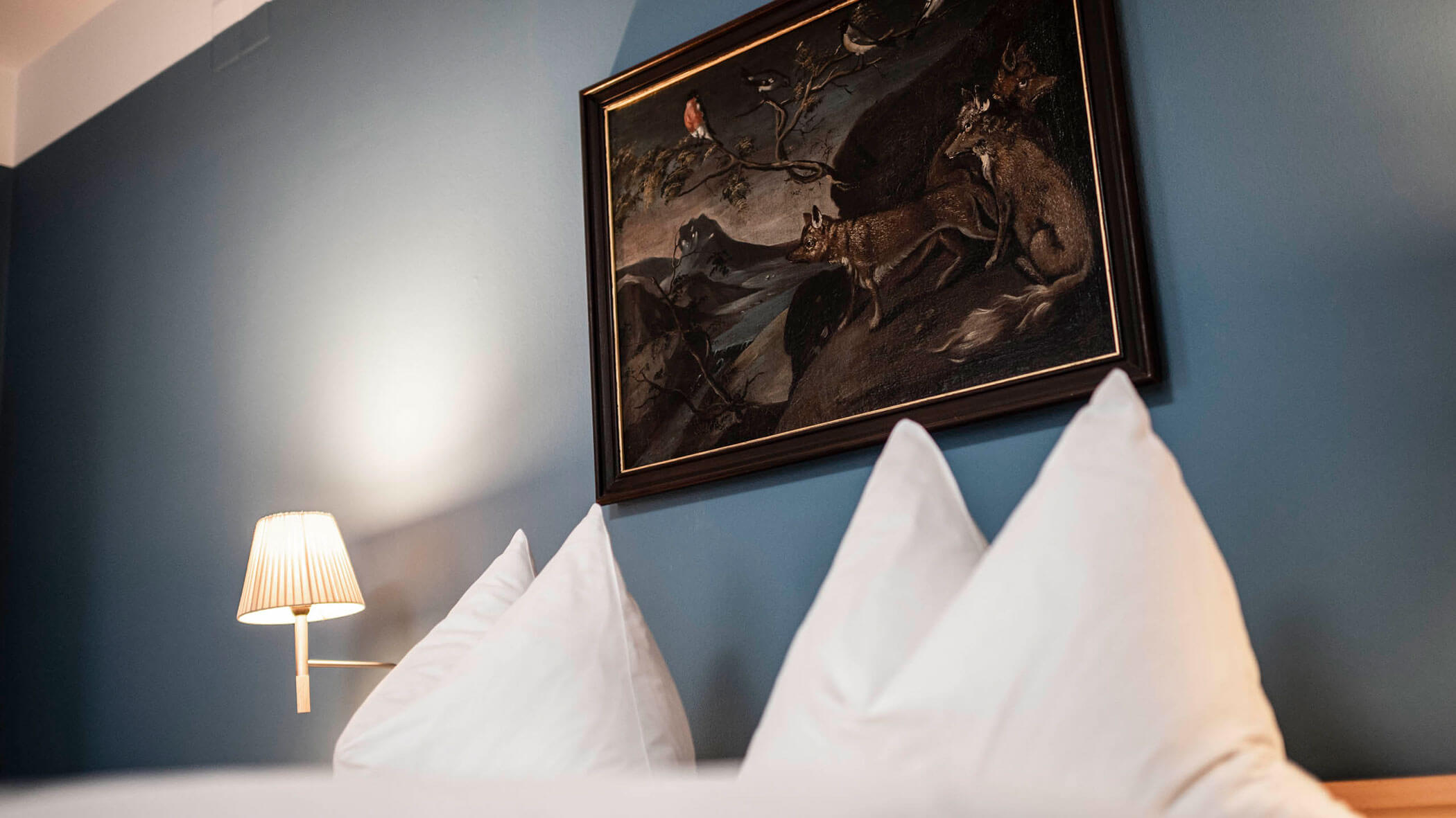 White pillows on a bed, a wall lamp, and a framed animal painting on a blue wall.