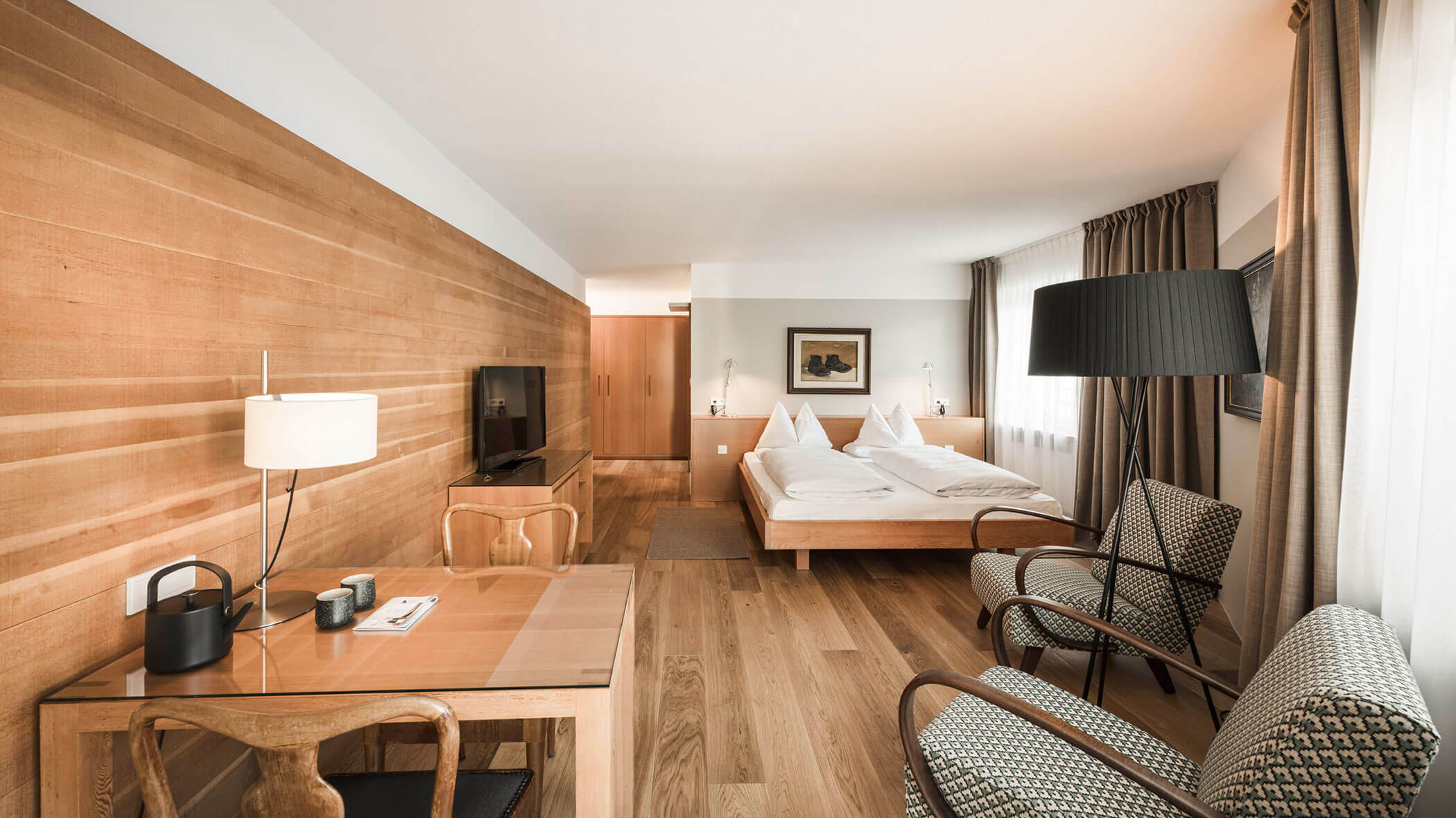 Modern hotel room with wooden floors, a double bed, desk, two chairs, TV, and large window with curtains.