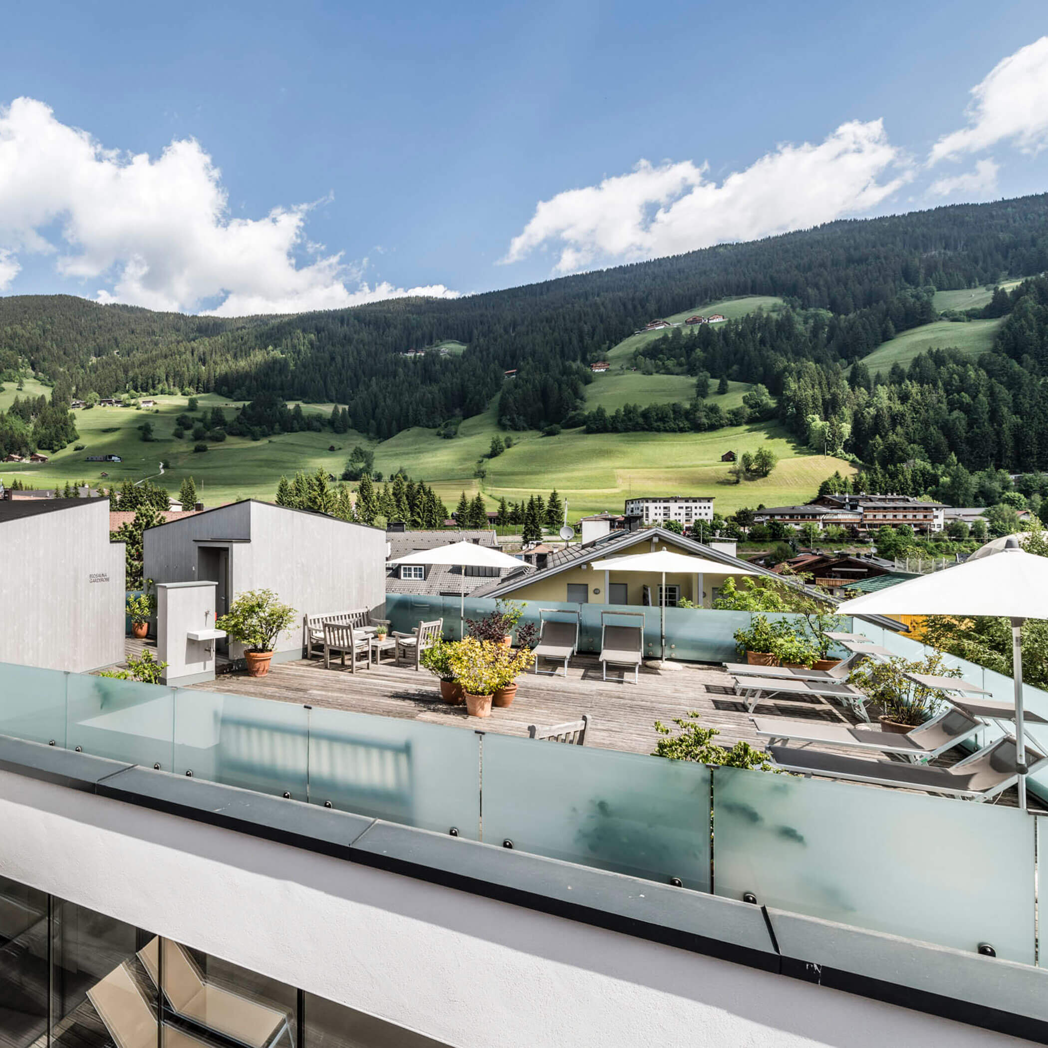 Rooftop terrace with lounge chairs and plants overlooking green hills and forested mountains under a blue sky.