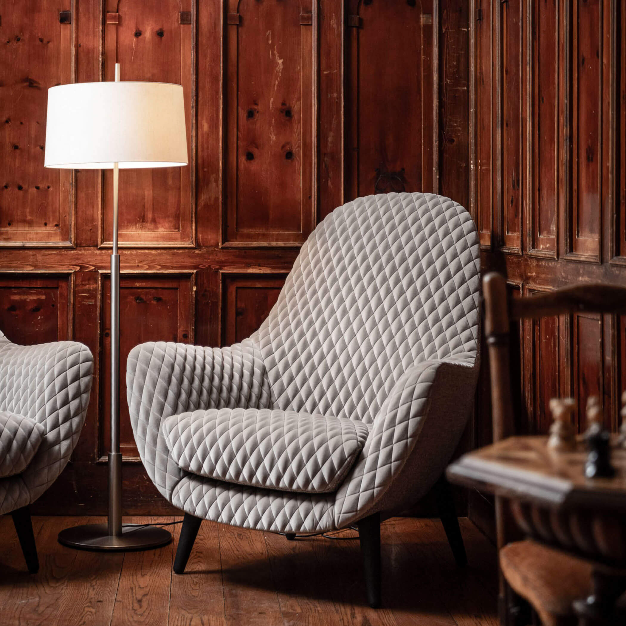 Quilted gray armchair beside a floor lamp in a cozy room with wooden paneled walls.