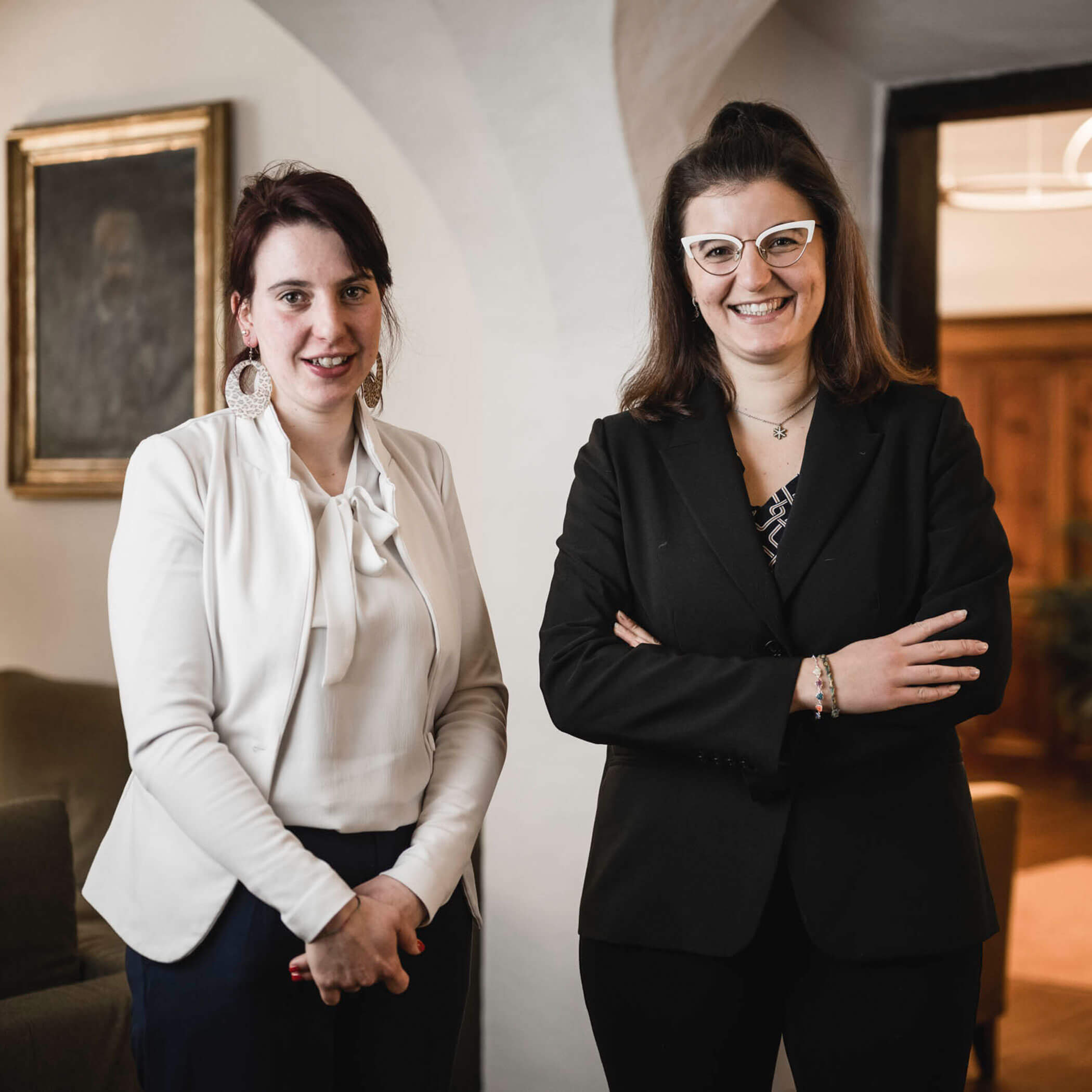 Two women in business attire smiling and standing indoors in a warmly lit room.