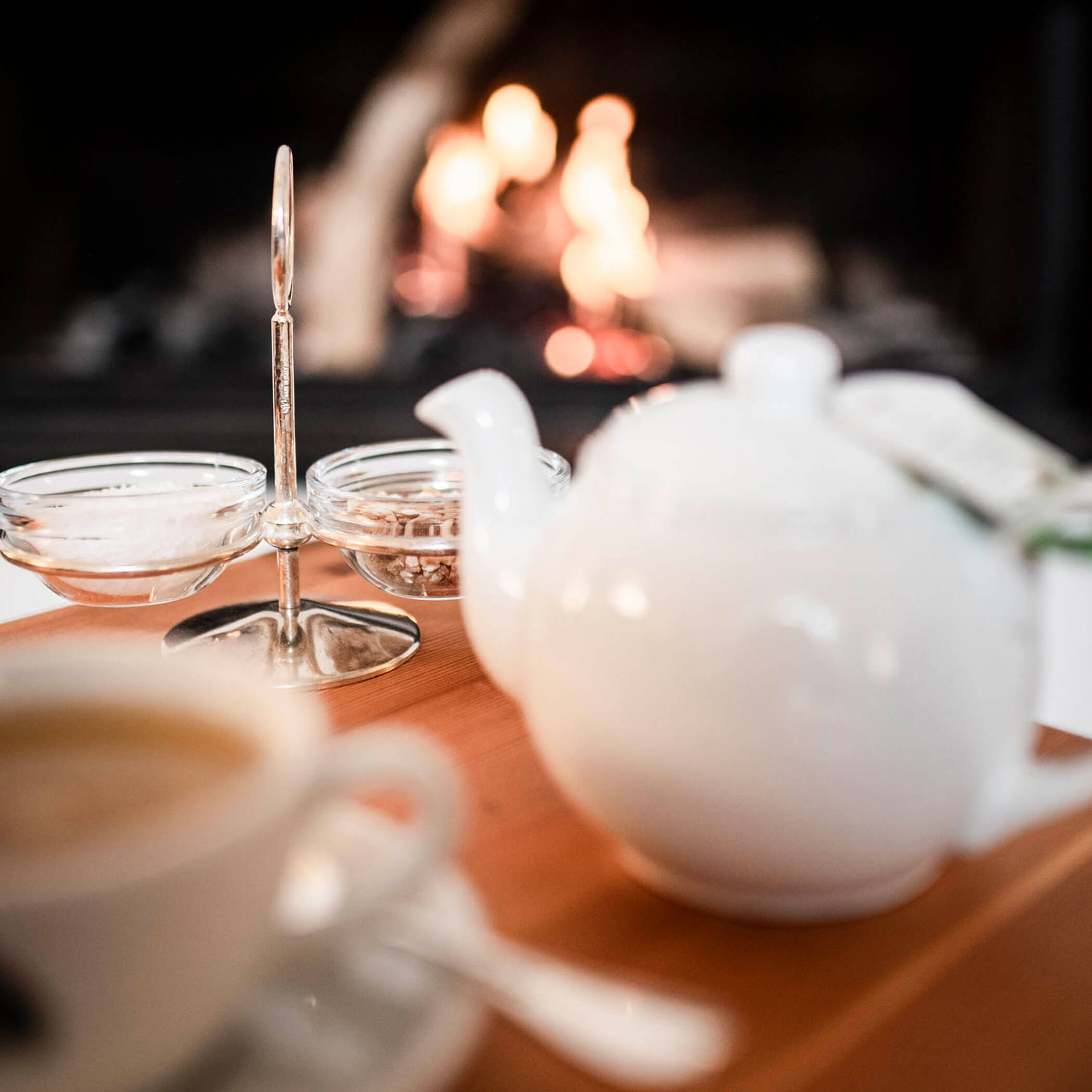 A teapot, teacup, and sugar bowls on a wooden table with a fire burning in the background.