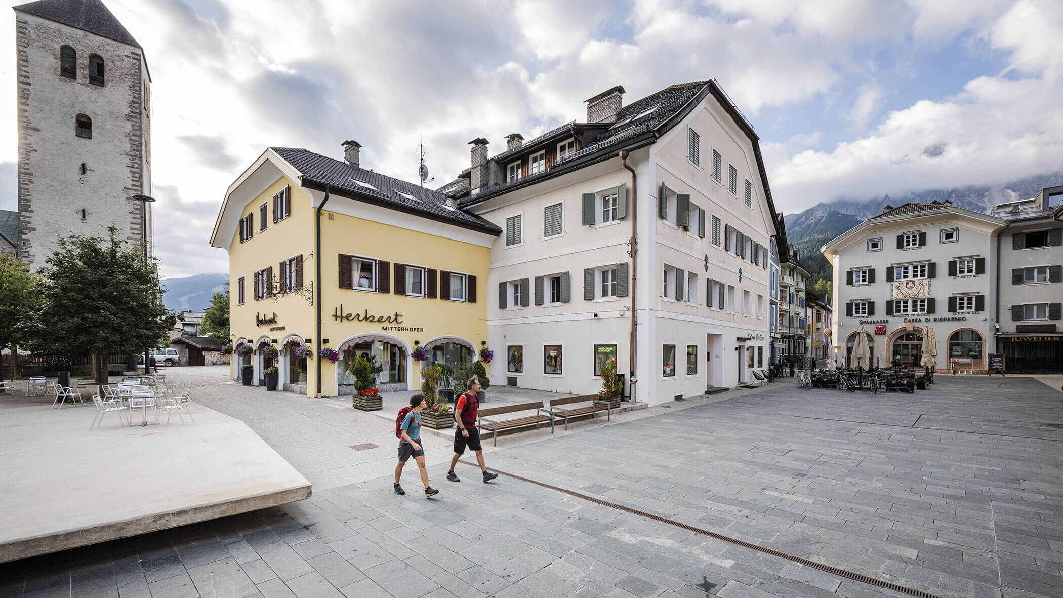 Two people walk across a quiet European square with historic buildings and mountains in the background.