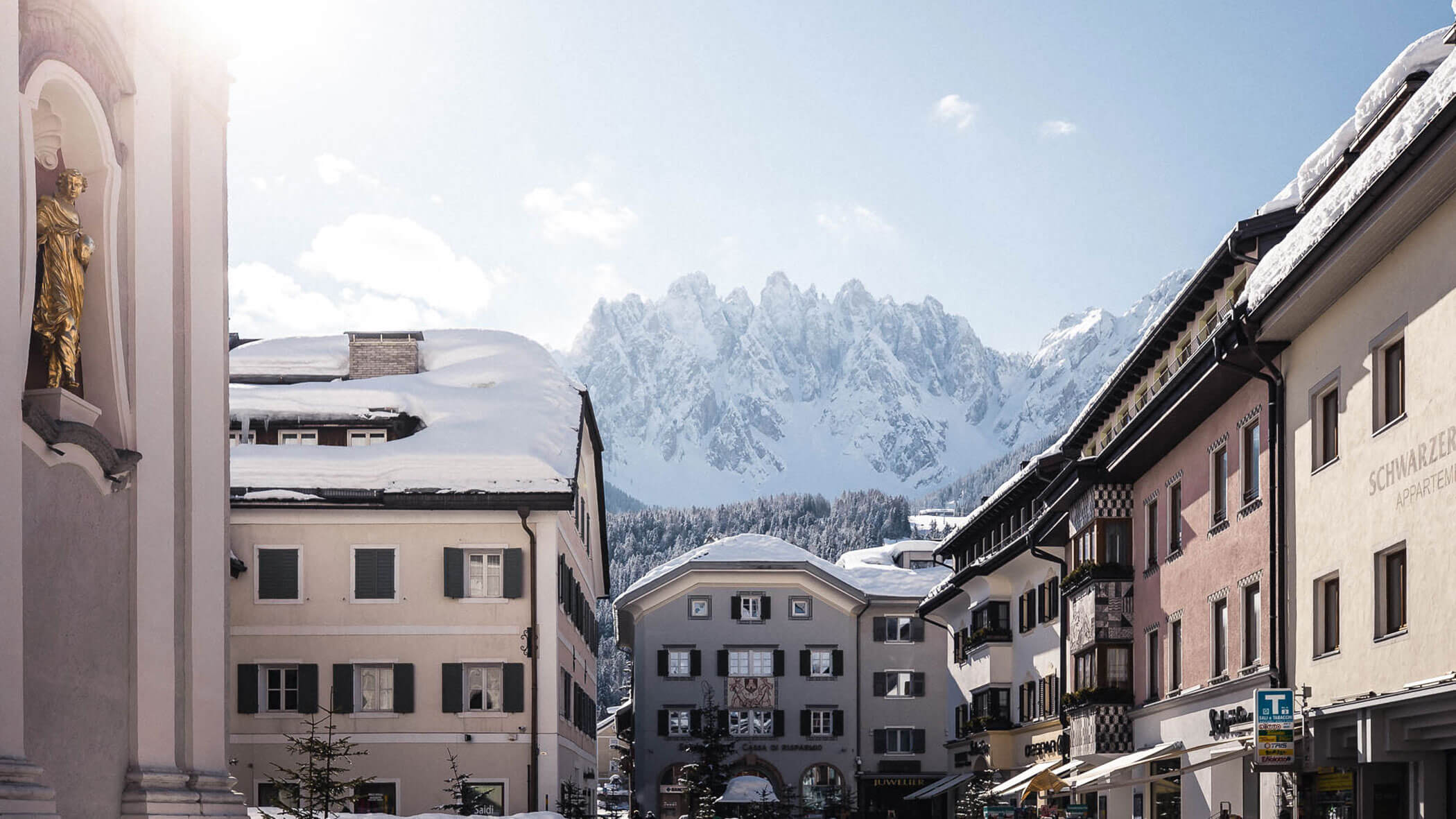 Snow-covered alpine village with tall buildings, mountain peaks in the background, and sunlight shining in.