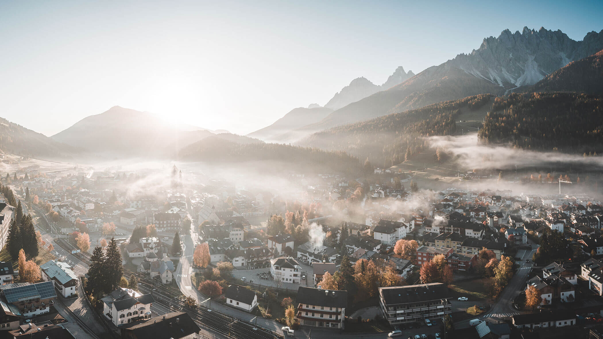 A foggy mountain town at sunrise, with autumn trees and mist rolling over rooftops and hills.