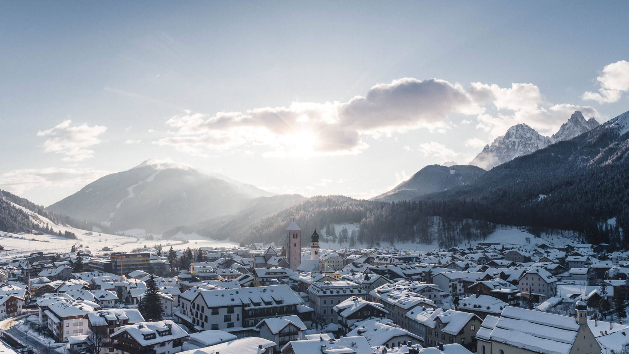 Snow-covered village with mountains in the background under a bright, partly cloudy sky at sunrise.