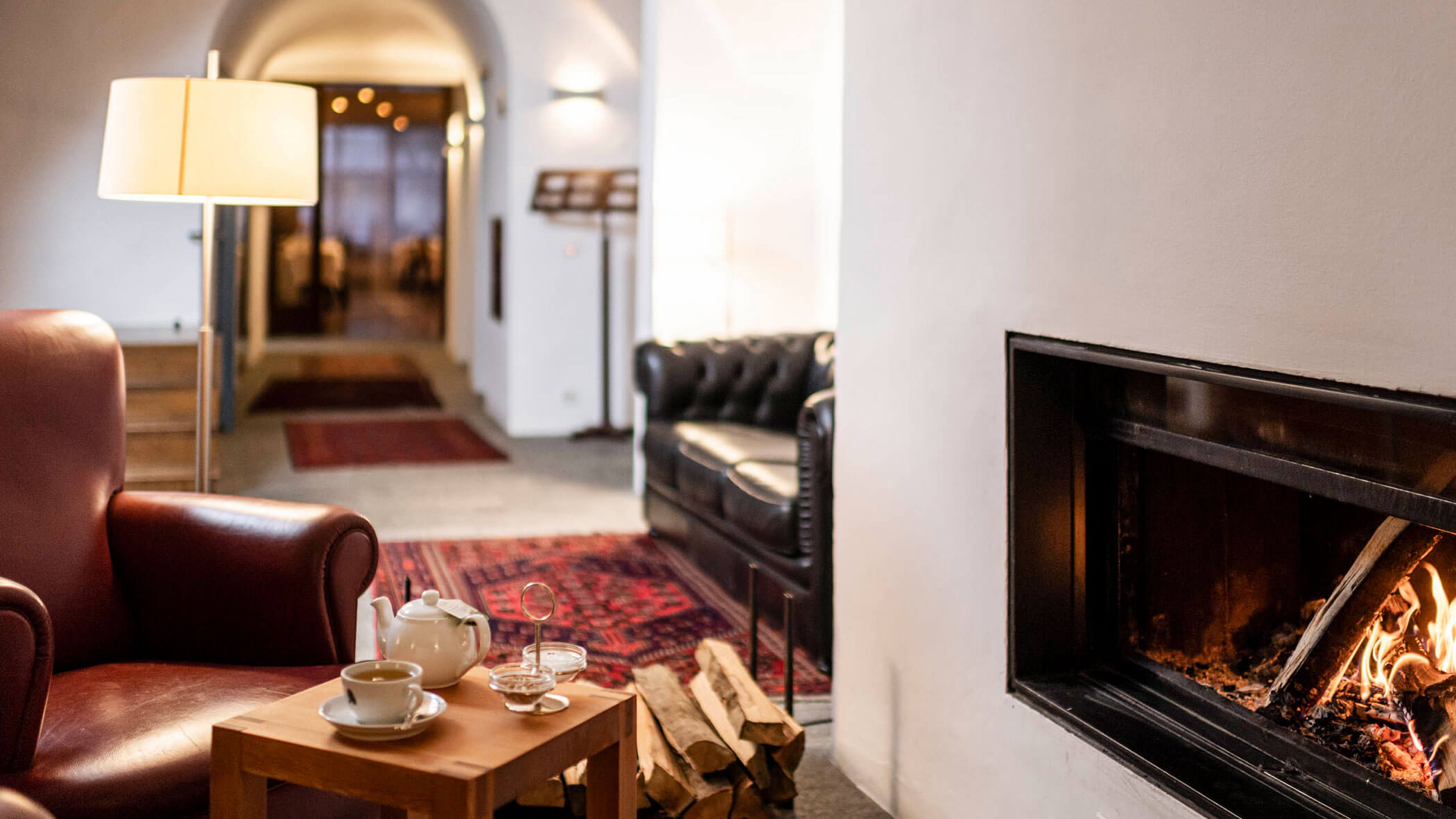 A cozy lounge with a lit fireplace, leather chair, tea set on a table, and a black sofa in the background.