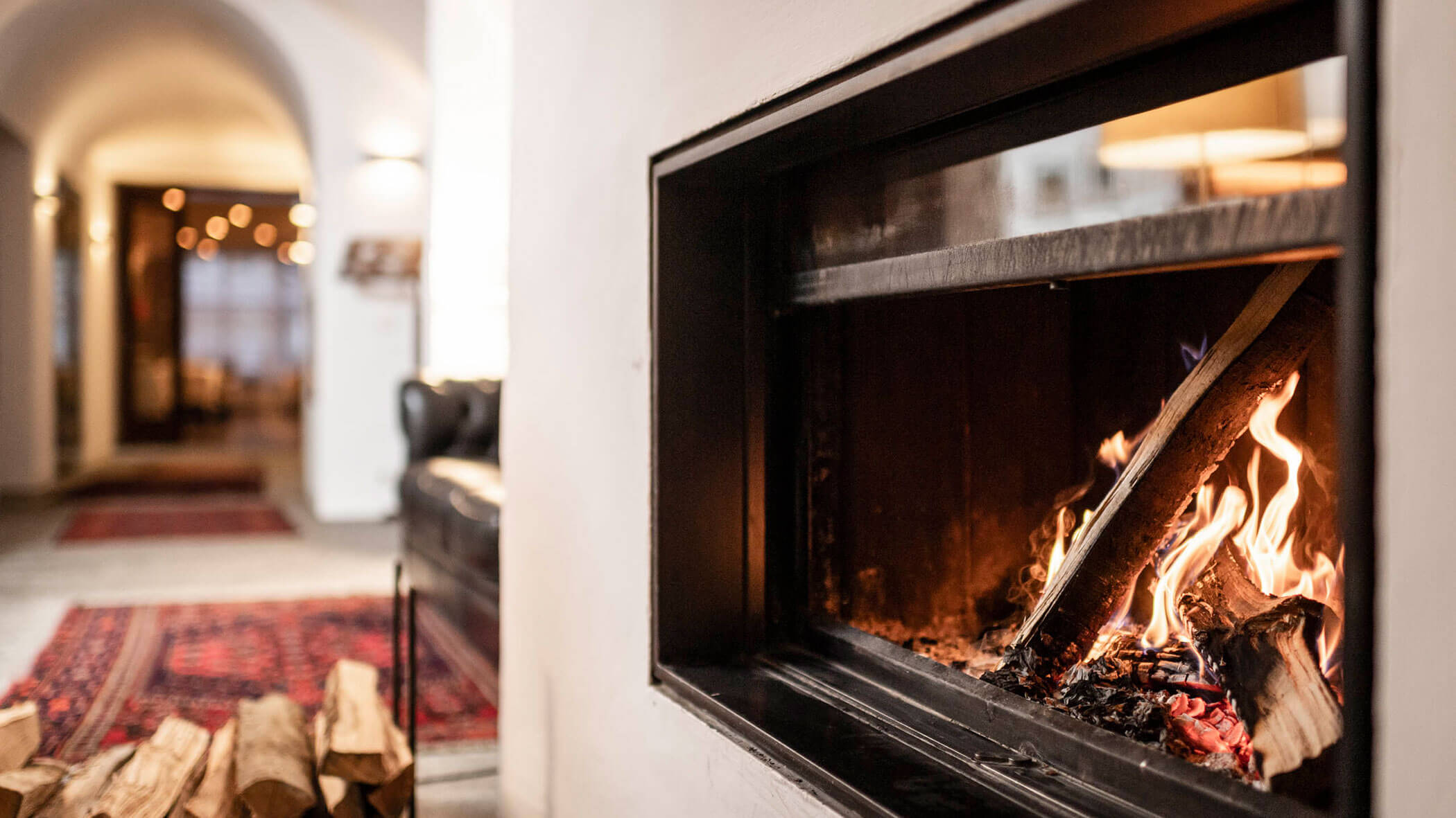 A cozy fireplace with burning logs in a warmly lit living room with rugs and a sofa in the background.