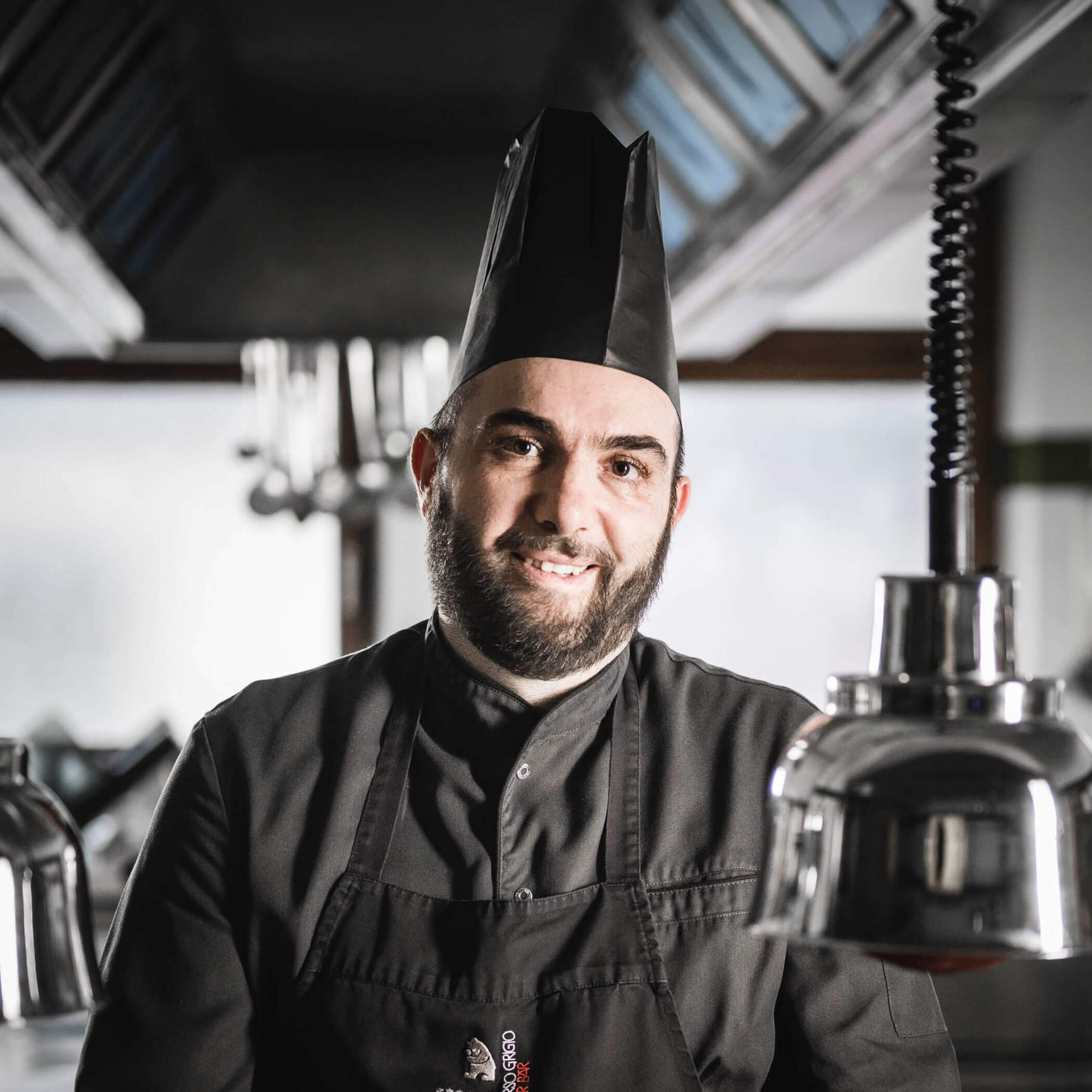 A smiling chef in a black uniform and hat stands in a professional kitchen.