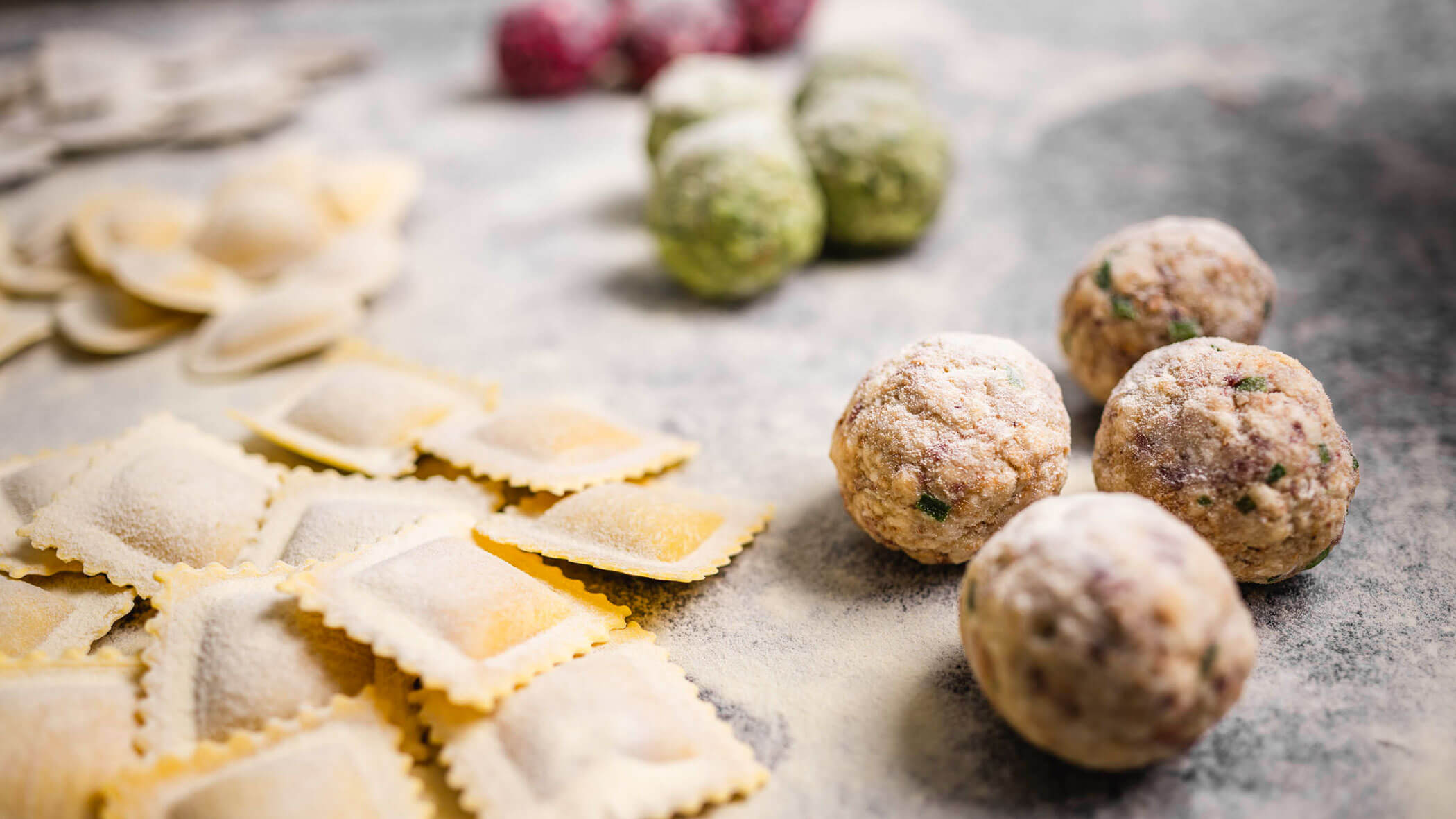 Fresh ravioli and uncooked meatballs dusted with flour on a countertop, with blurred ingredients in the background.