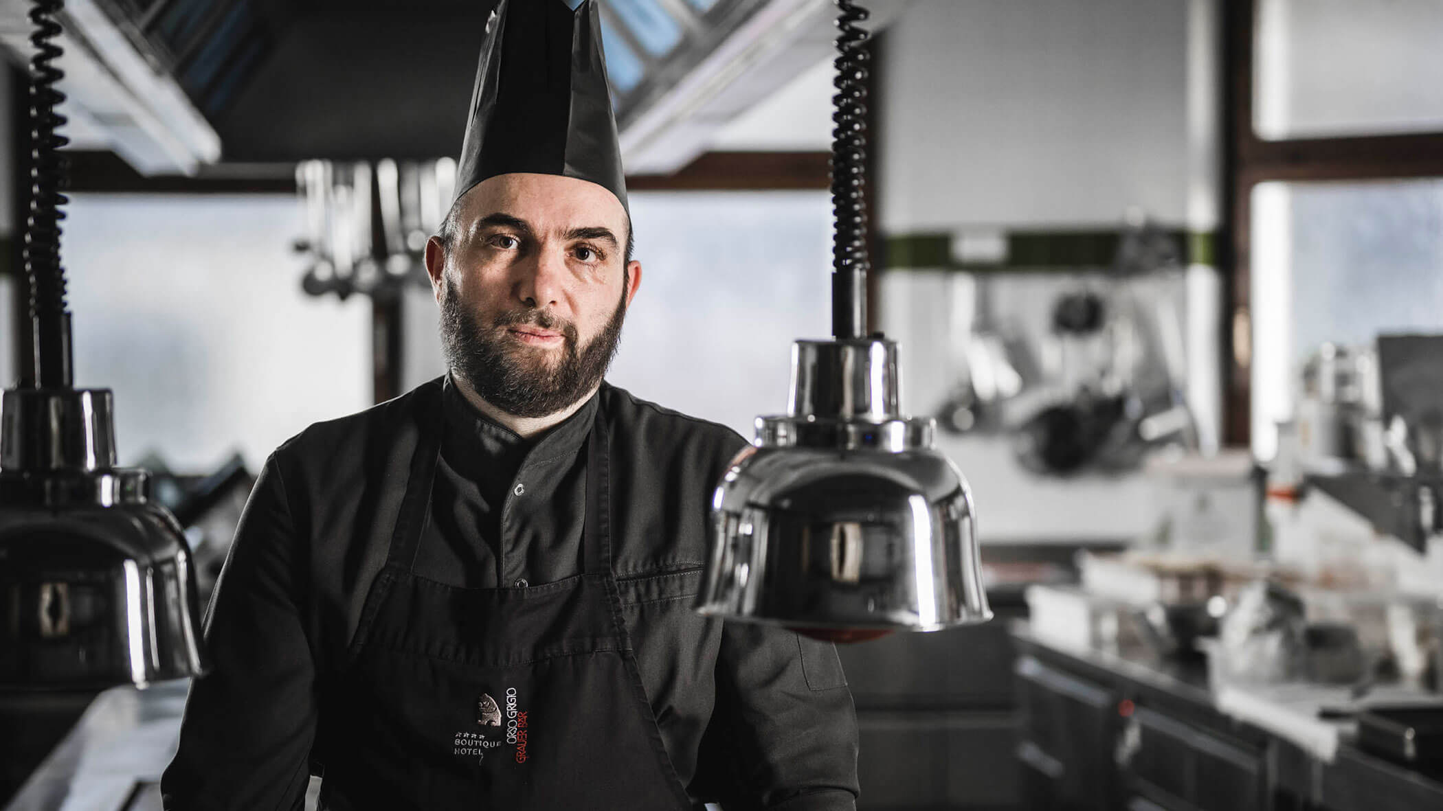 A chef in a black uniform and hat stands in a professional kitchen, looking at the camera.