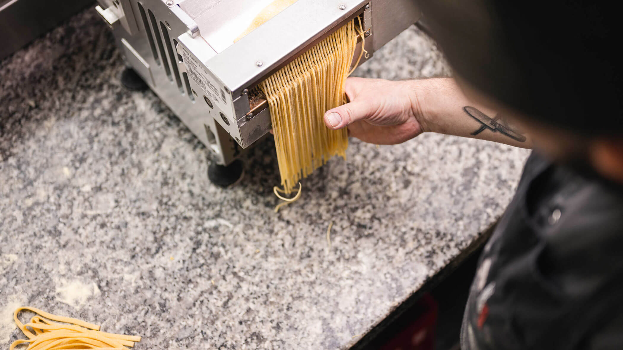 Person using a pasta machine to make fresh noodles on a speckled countertop.