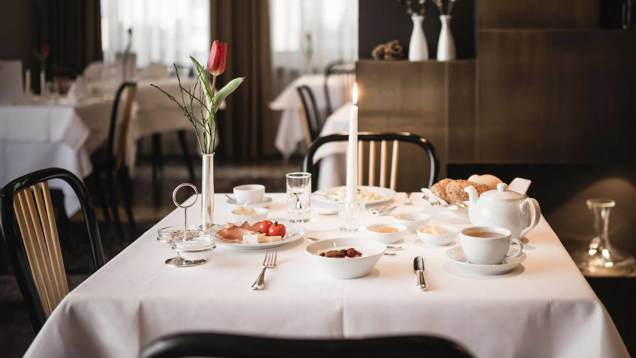 Elegant breakfast table set with dishes, a teapot, a lit candle, and a single red tulip in a vase.