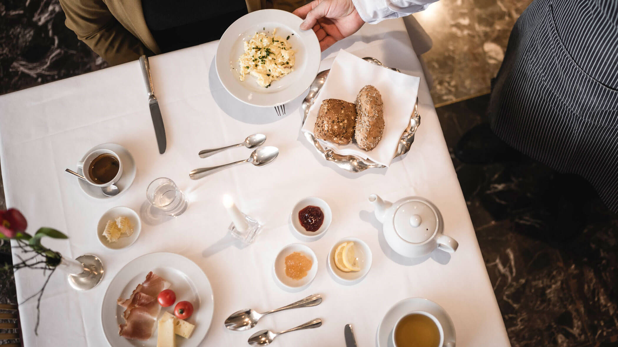 A server hands a plate of scrambled eggs to a seated person at a breakfast table set with bread, tea, and spreads.