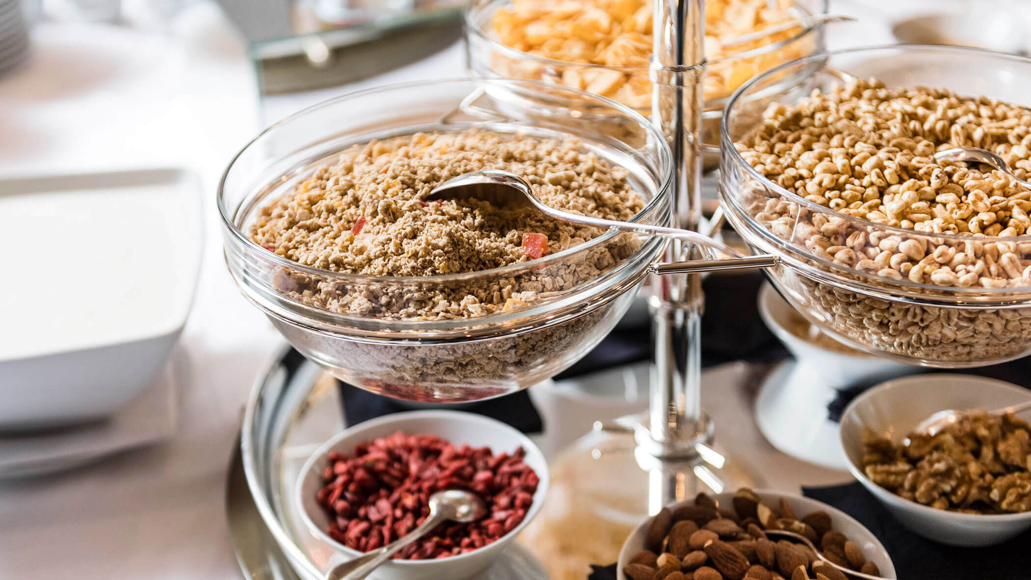 Glass bowls filled with different cereals, nuts, and seeds on a breakfast buffet with serving spoons.
