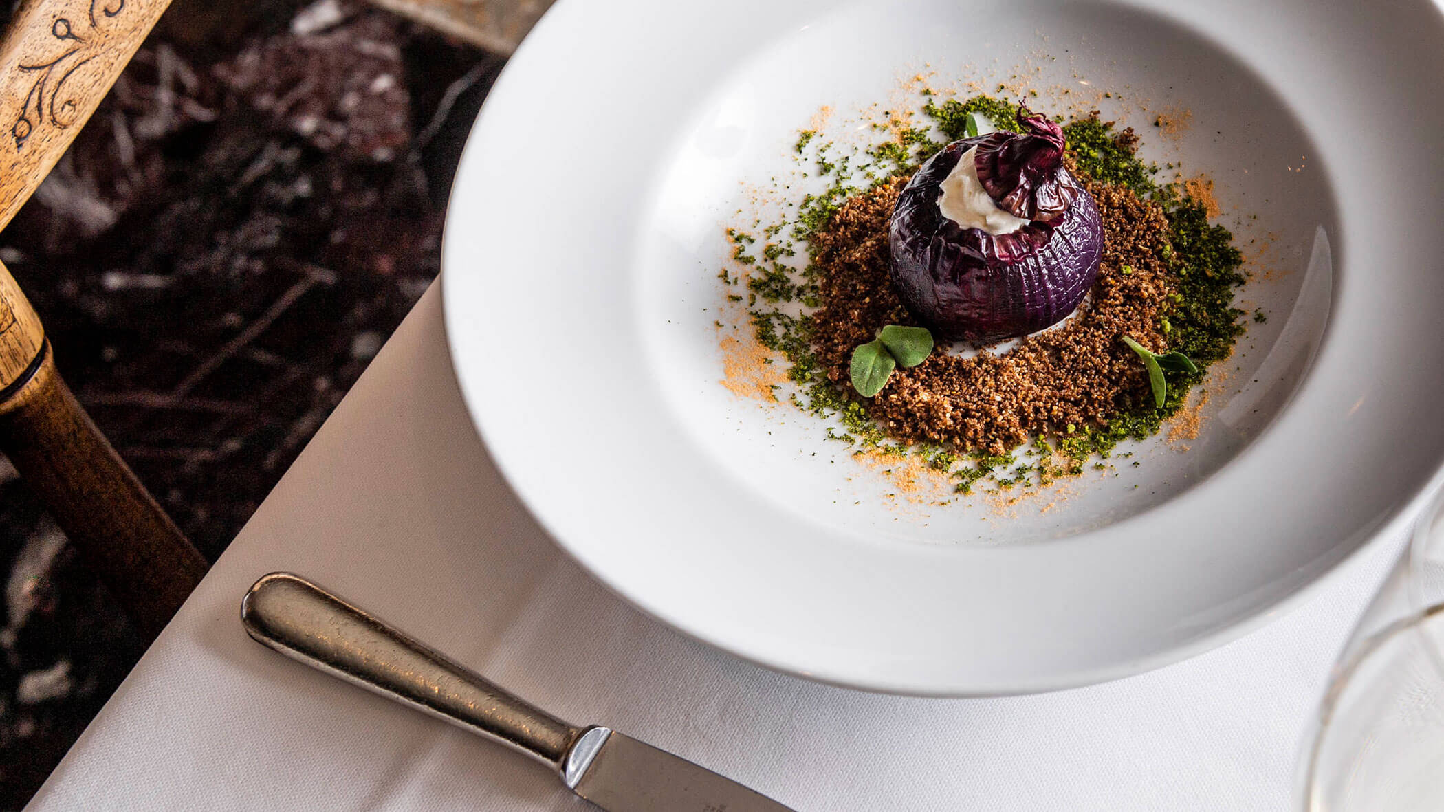 A gourmet purple onion dish garnished with herbs and crumbs on a white plate, set on a table with a knife.