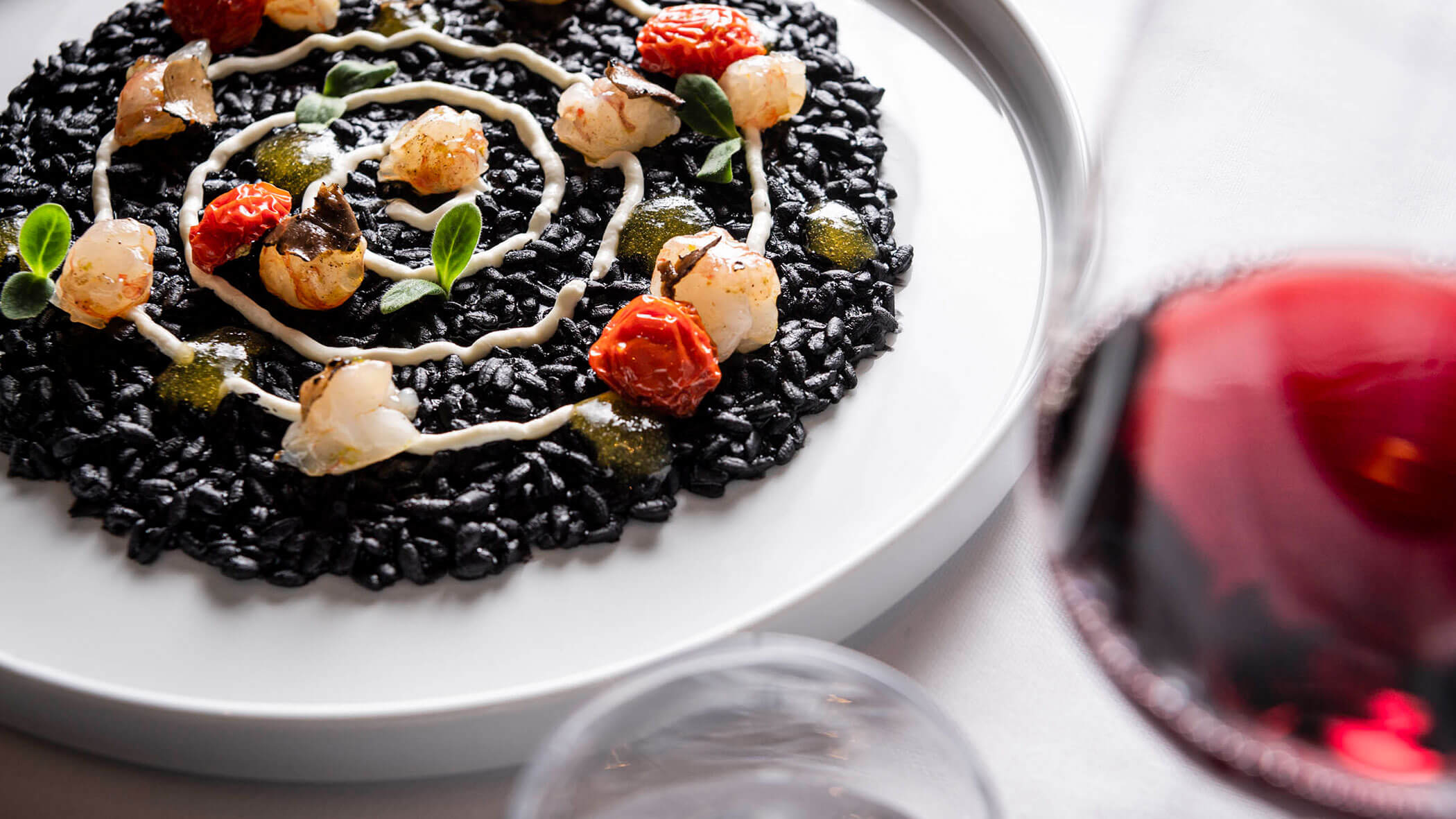 Black risotto garnished with seafood, tomatoes, and herbs, artfully plated with a glass of red wine nearby.