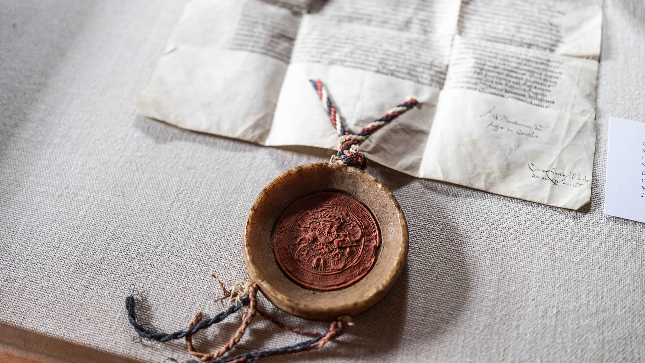A wax seal with multicolored cords lies in front of an old, partially folded document with handwritten text.