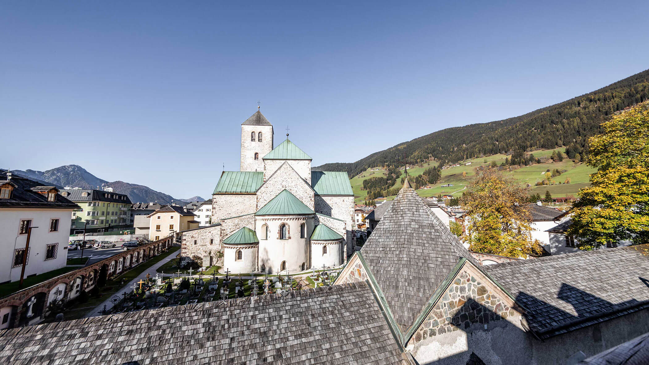 A historic church with green roofs surrounded by a village and mountains under a clear blue sky.