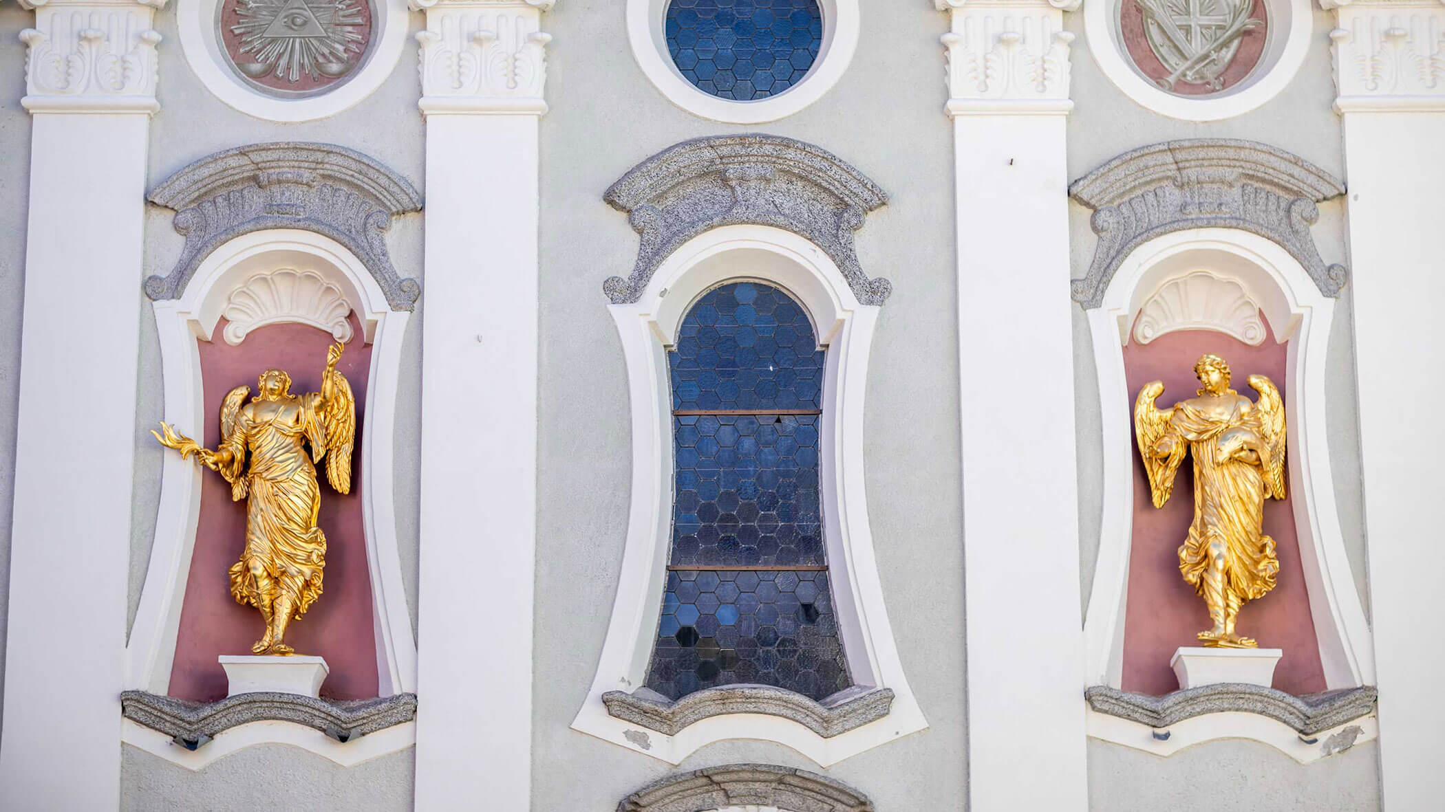 Two golden angel statues flank a blue-tinted arched window on an ornate building facade.