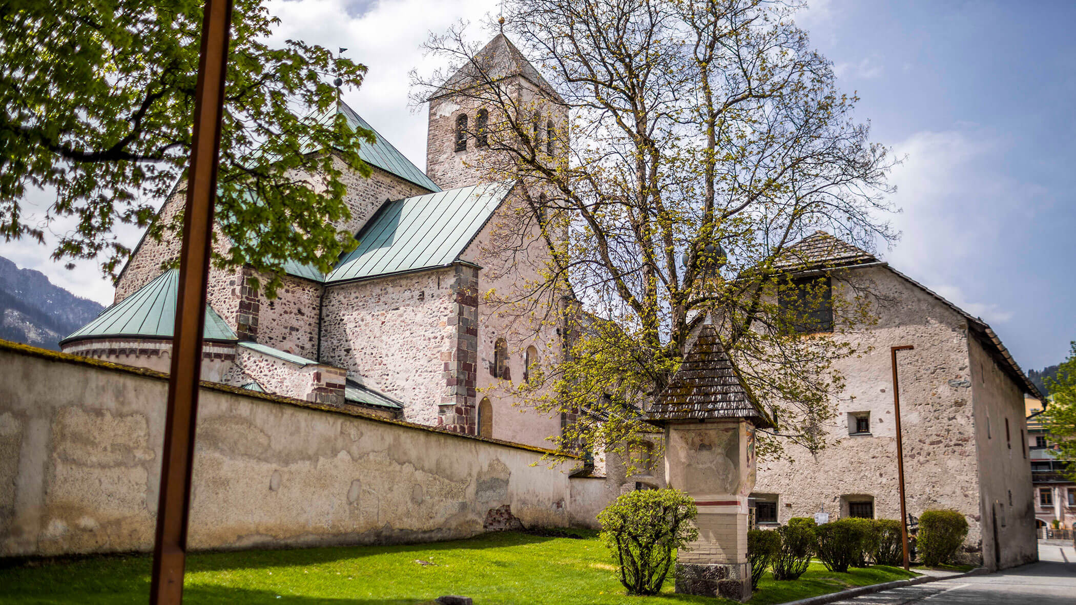 Chiesa in pietra con tetto verde, torre e albero, circondata da un muro di pietra e da un prato verde in una giornata di sole.