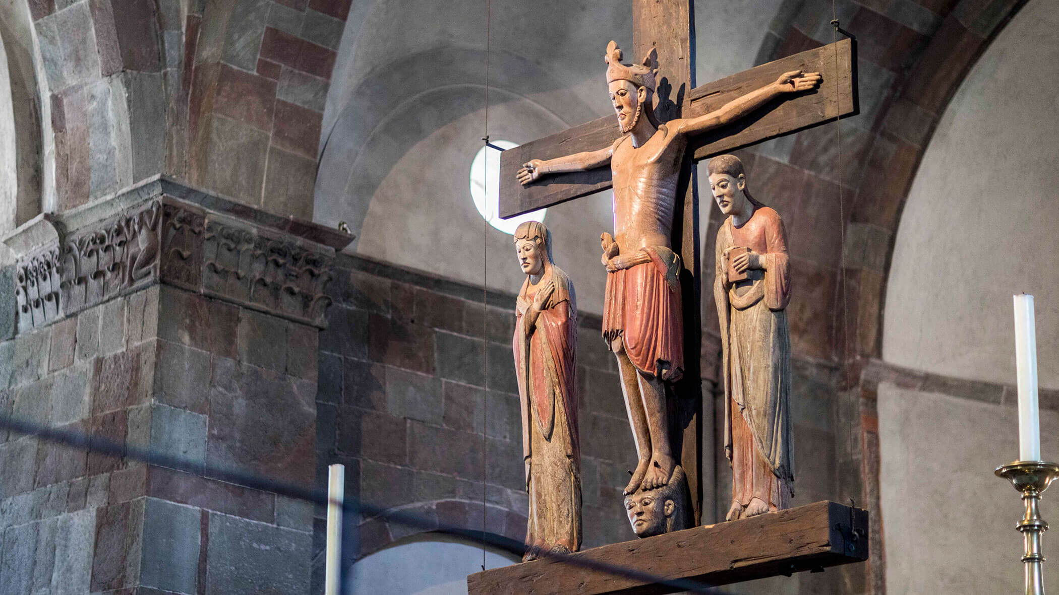 Wooden crucifix with three carved religious figures inside a stone church with arched ceilings and columns.